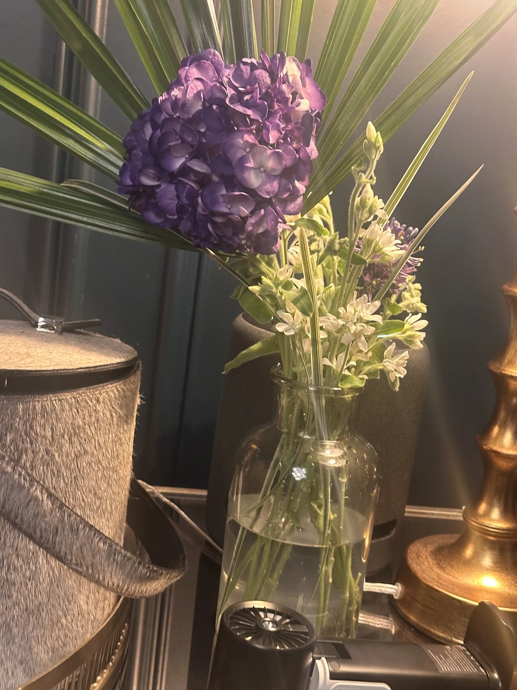 Inside a kitchen, a clear glass vase with purple and white flowers sits on a countertop next to a coffee machine and a decorative object. The background includes a cabinet and a metallic wall.
