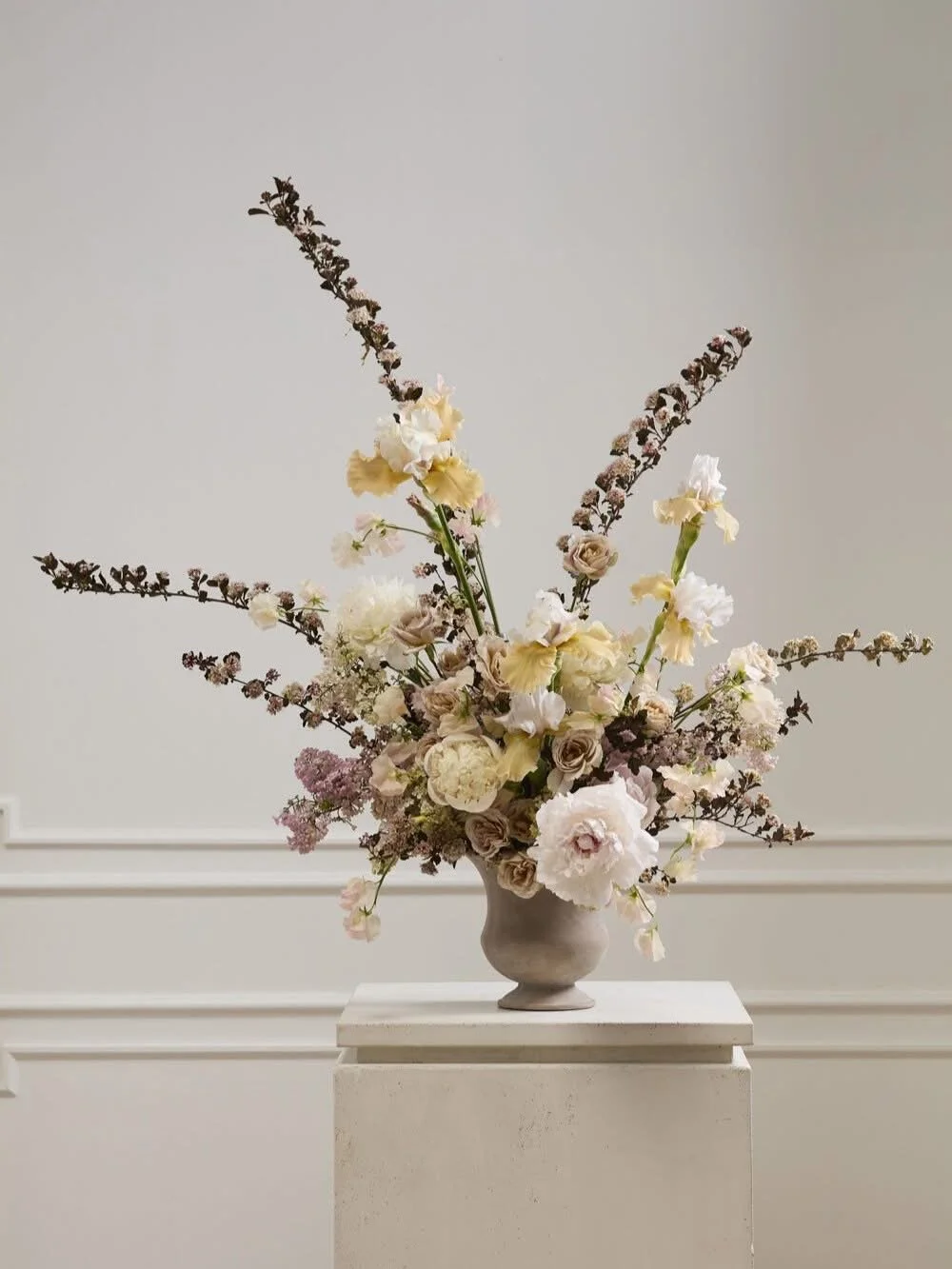 A large floral arrangement in a gray ceramic vase, featuring white, cream, and light pink flowers, with some purple and dark accent flowers, on a white pedestal against a plain light gray wall.