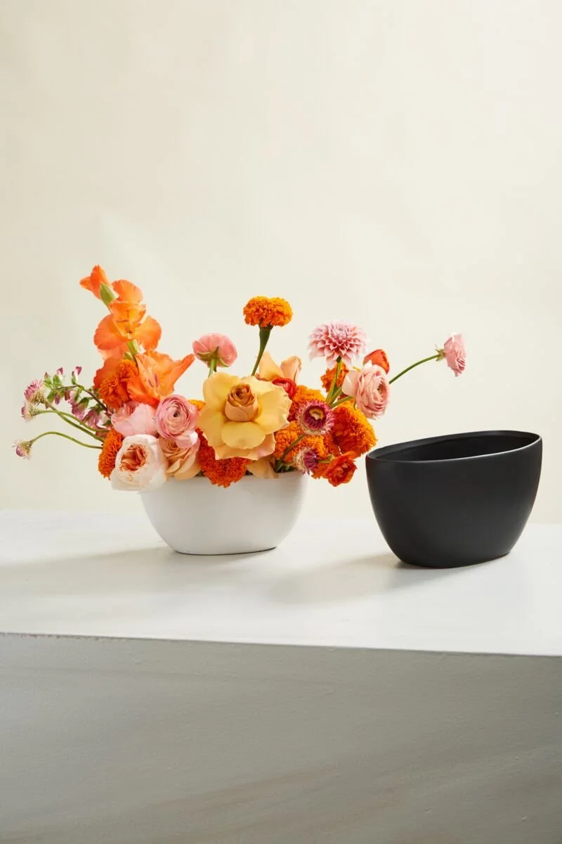 A white bowl with a colorful bouquet of pink, orange, yellow, and peach flowers next to a plain black bowl on a white surface against a light-colored wall.