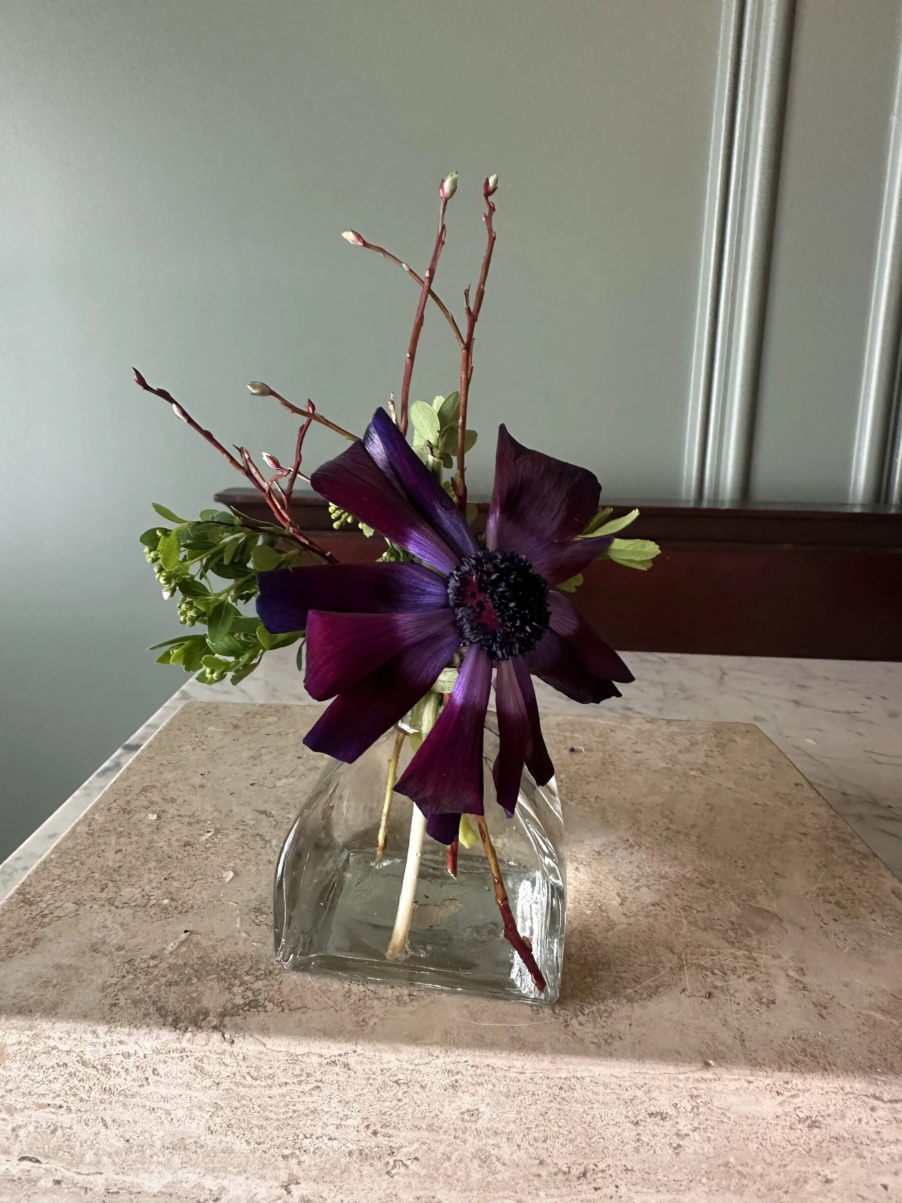 A floral arrangement in a clear glass vase with purple and green flowers and branches, placed on a marble countertop.