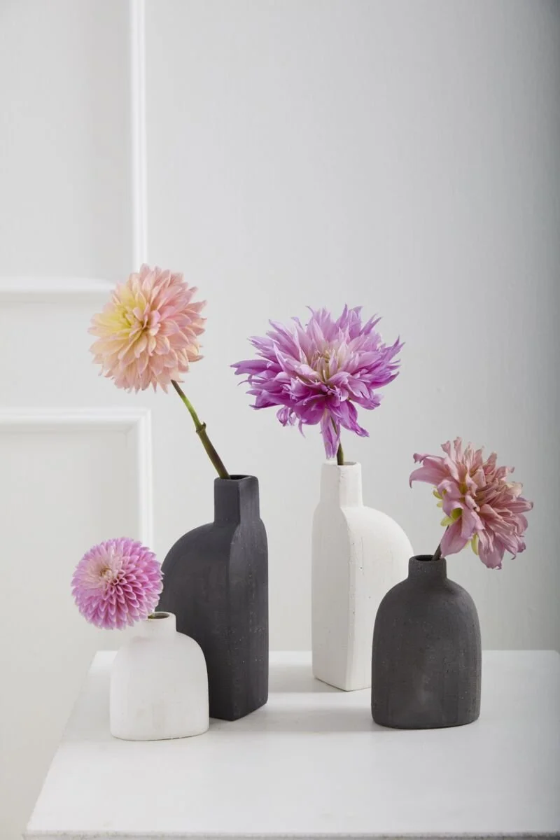 Five vases with artificial flowers arranged on a white table against a white wall, featuring pink and purple dahlias in black and white vases.