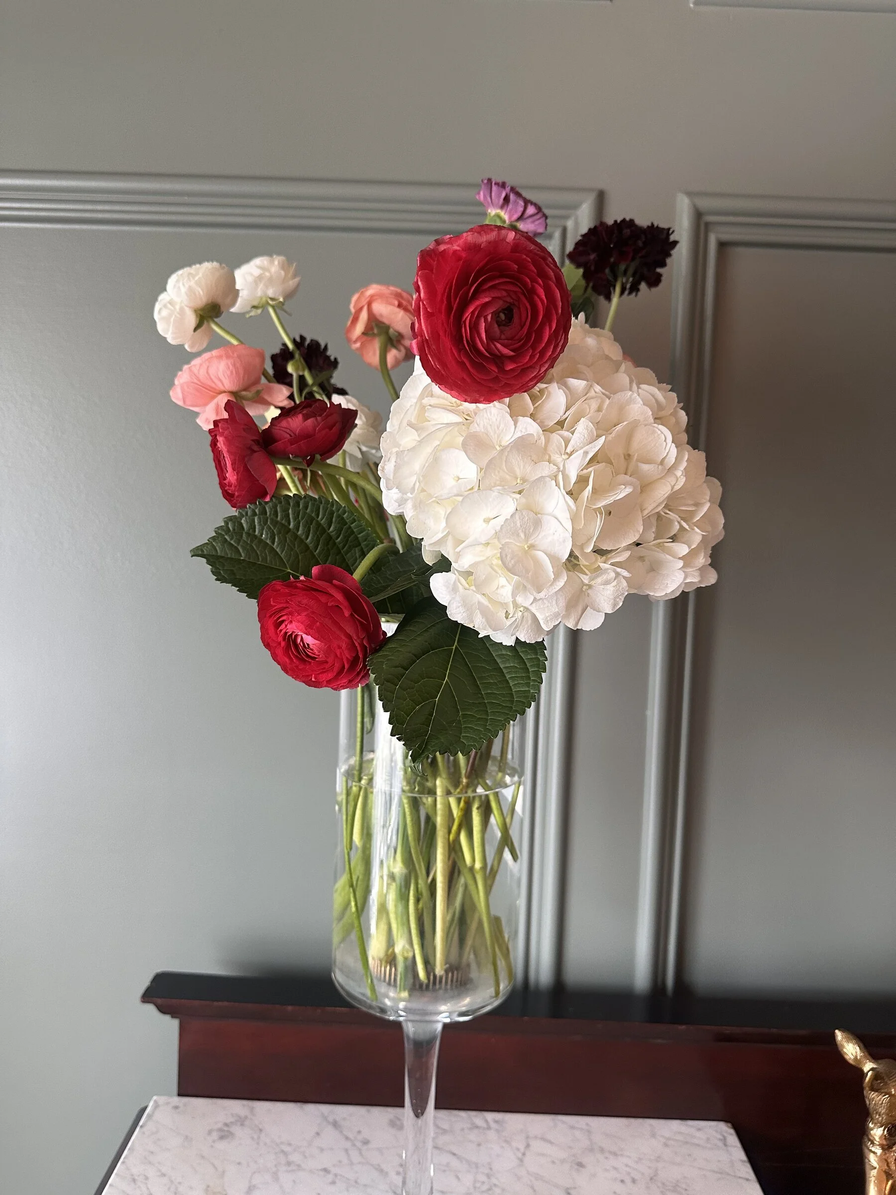 A tall glass vase filled with white hydrangea, pink, red, and dark purple ranunculus flowers, and green leaves on a marble surface in front of a gray wall with decorative molding.