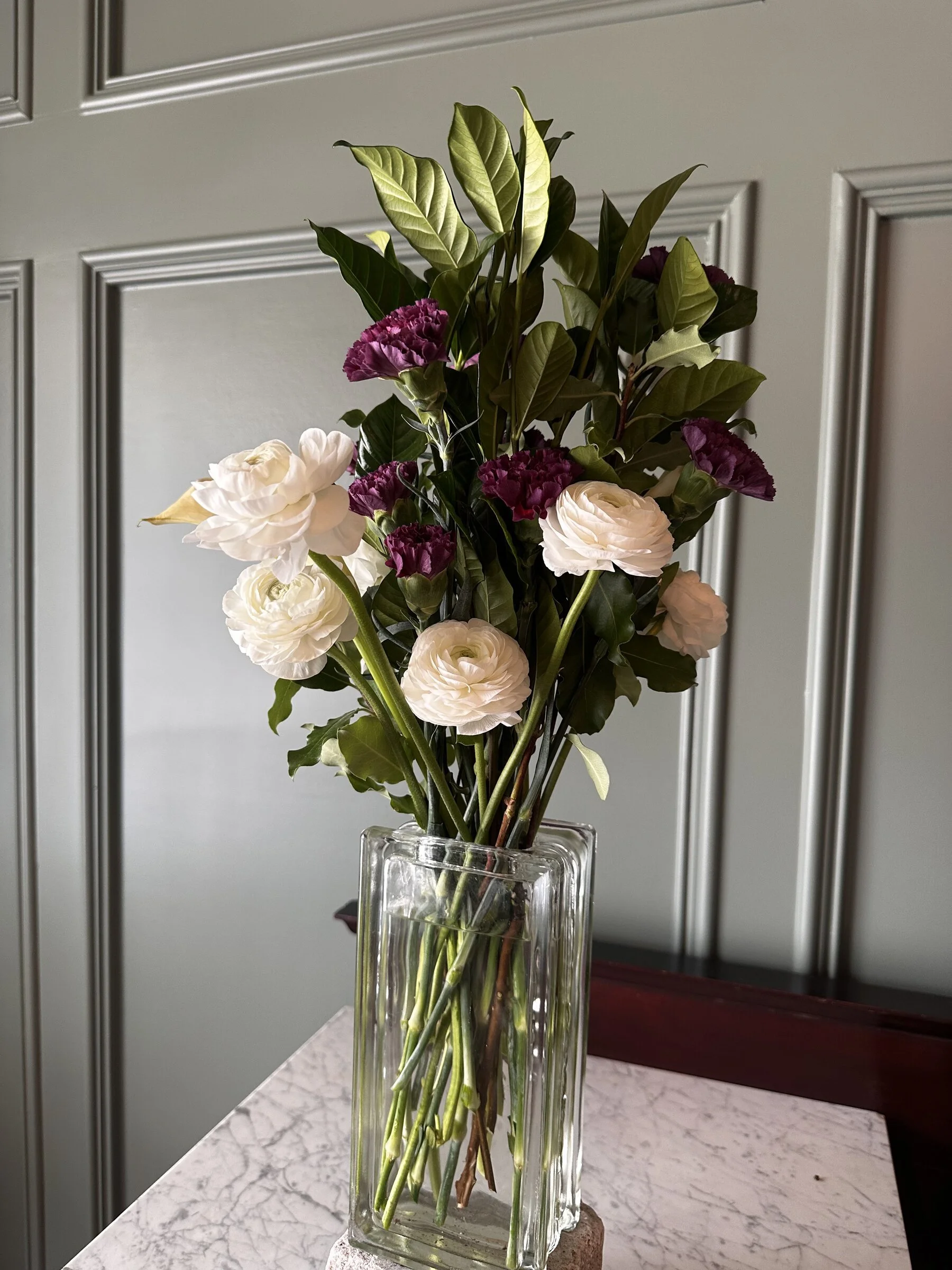 A bouquet of white and purple flowers with green leaves in a tall glass vase on a white marble table, with gray paneled wall in the background.