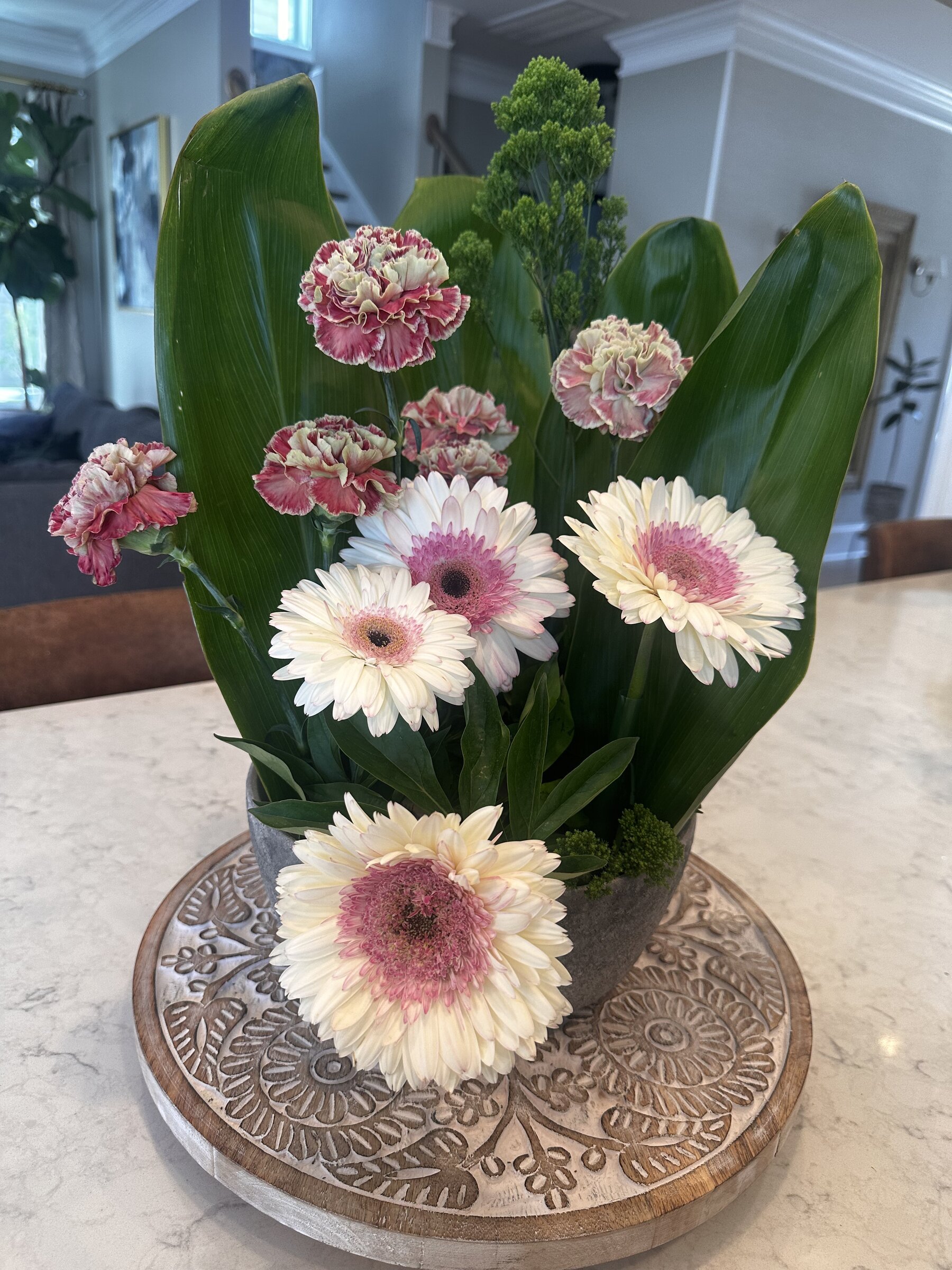A floral arrangement with pink and white carnations, white gerbera daisies, and green foliage in a gray ceramic pot, placed on a decorative wooden tray on a kitchen counter.