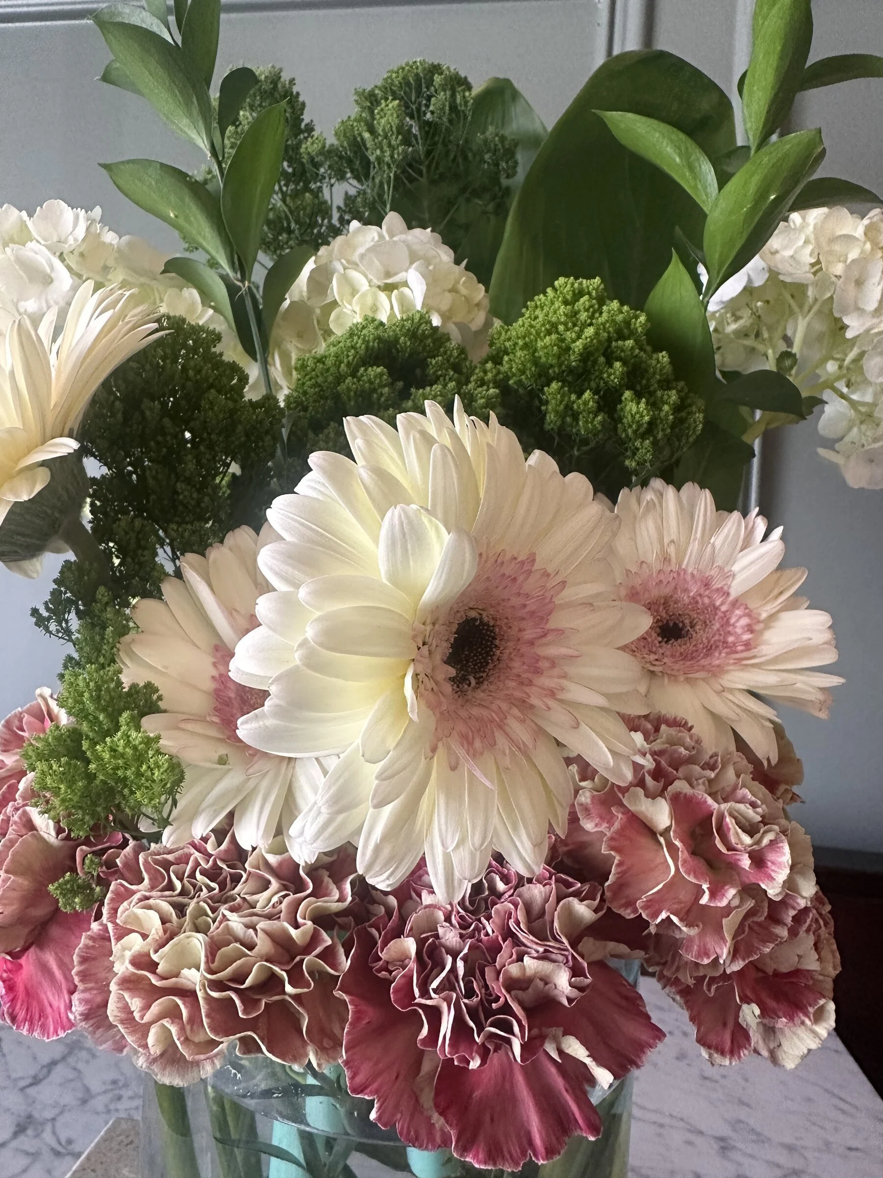A bouquet of mixed flowers in a vase, including white gerbera daisies, pink/yellow carnations, green leafy foliage, and other greenery.