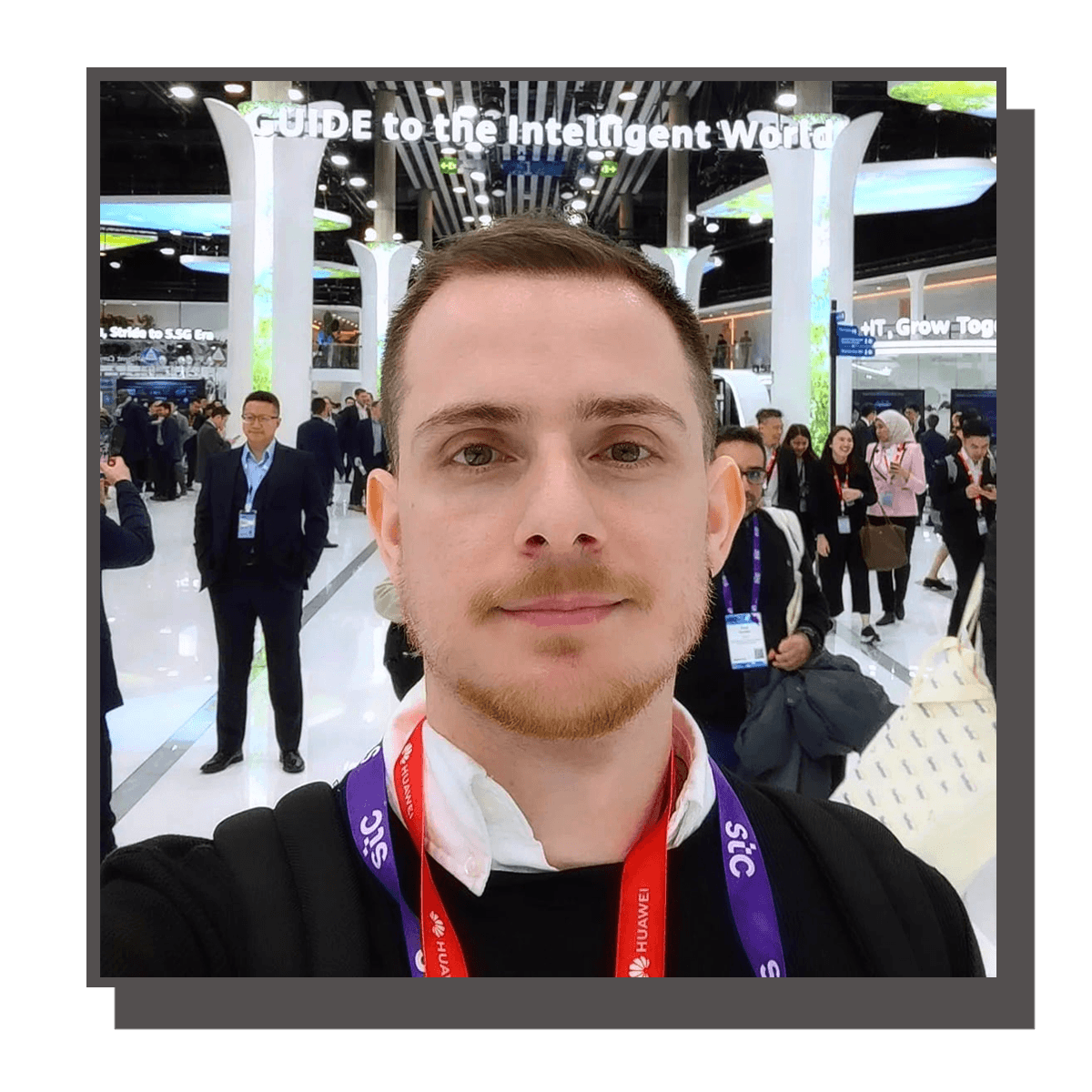 A man taking a selfie at a technology conference with many people in the background, large digital displays, and signs including "Guide to the Intelligent World".
