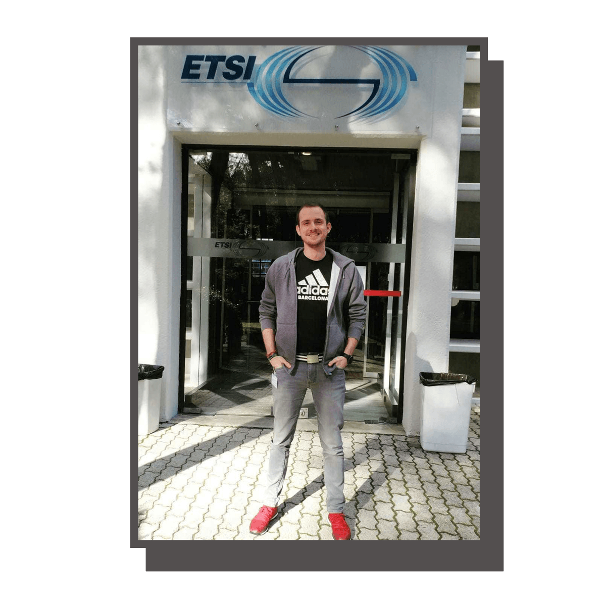 A young man standing outside the entrance of an ETSI building, smiling and wearing casual clothes including a black Adidas Barcelona T-shirt, gray hoodie, and red shoes. He is standing on a paved sidewalk.