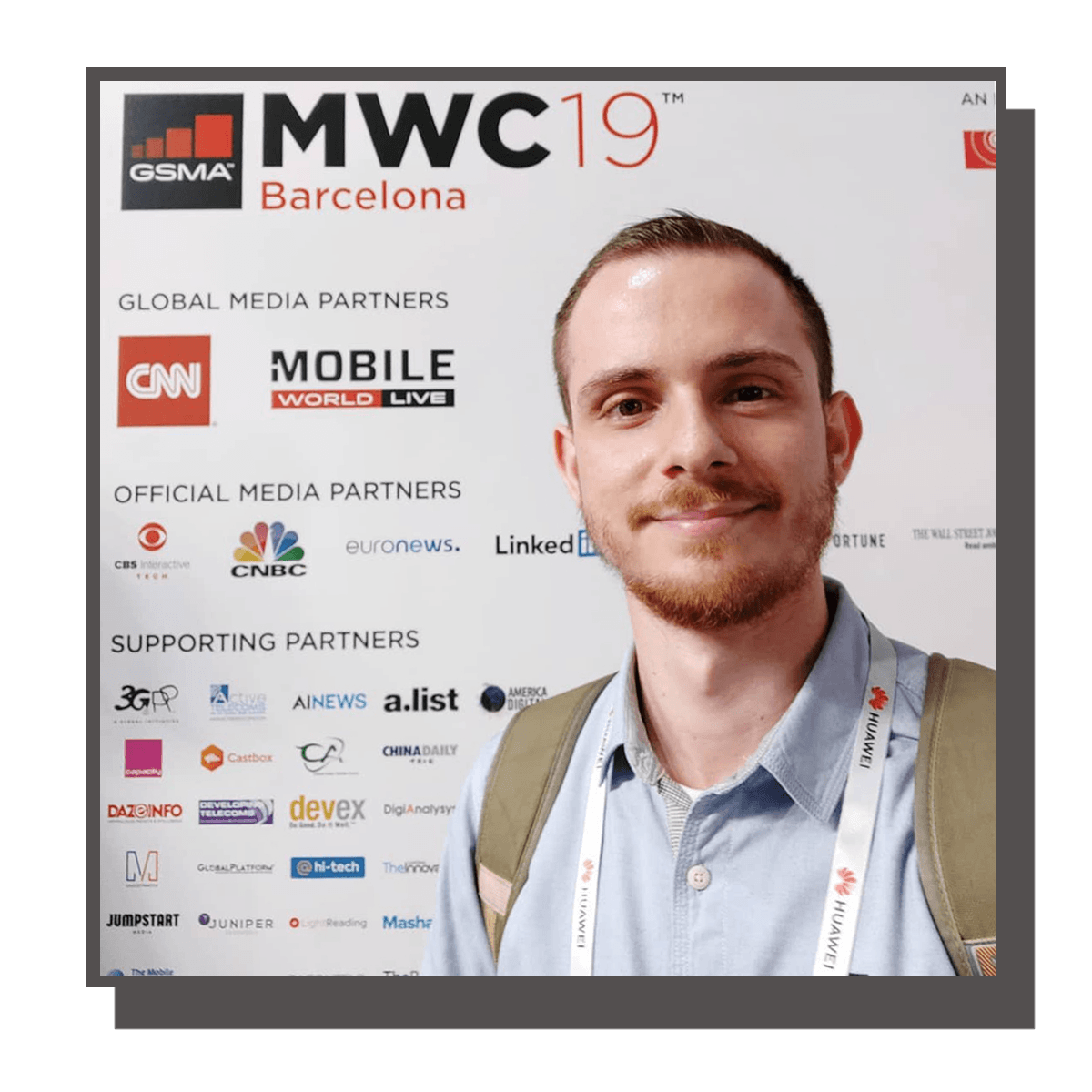 Young man with a light beard and short hair taking a selfie at the Mobile World Congress 2019 in Barcelona, standing in front of a backdrop with logos of media partners, including CNN, CNBC, LinkedIn, and others. He is smiling, wearing a light blue shirt, and has a badge and backpack with Huawei branding.
