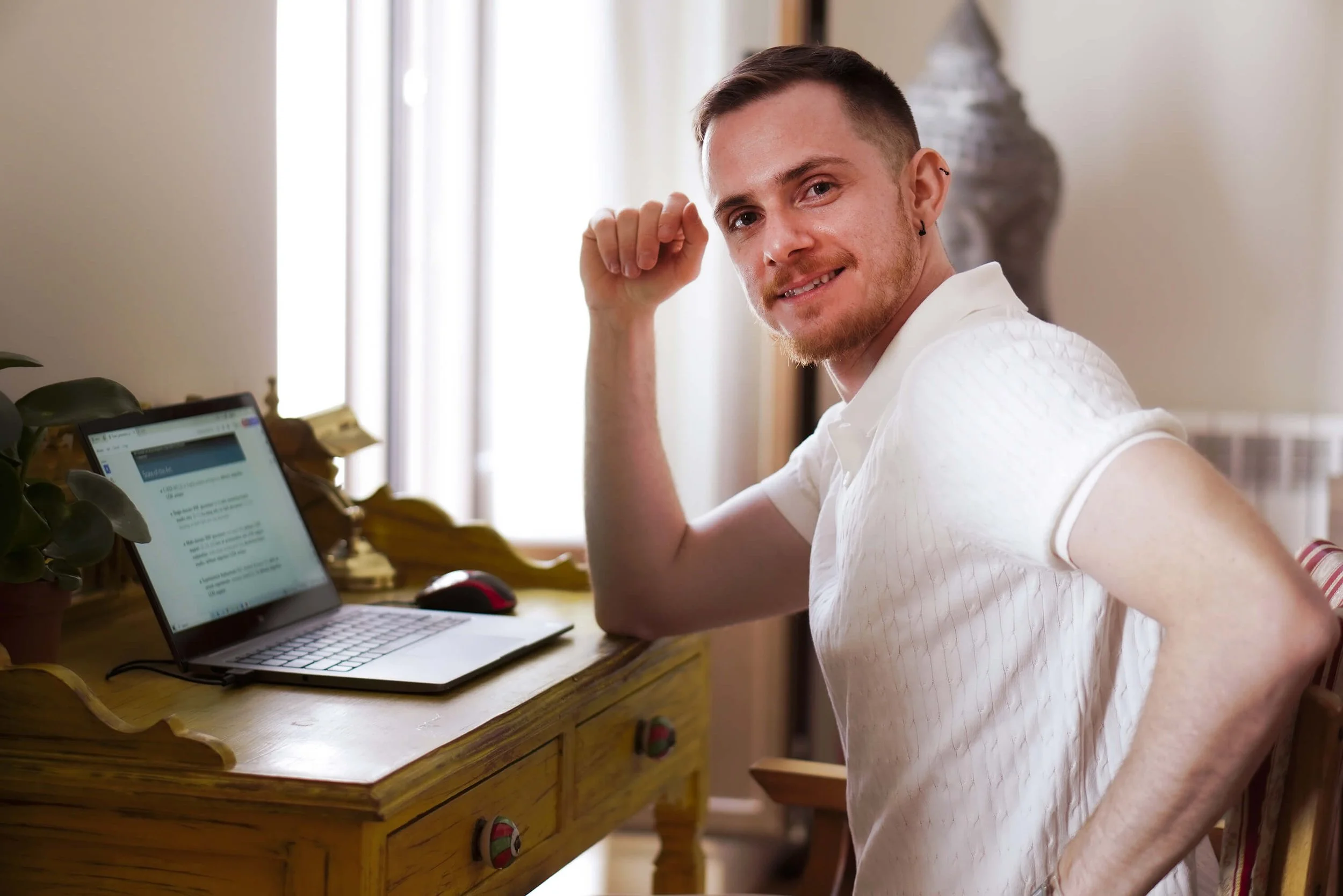 A young man with short brown hair, a beard, and a black earring is smiling at the camera while sitting at a wooden desk. He is wearing a white shirt and sitting in a well-lit room with beige walls, beside a laptop, a mouse, and some papers.