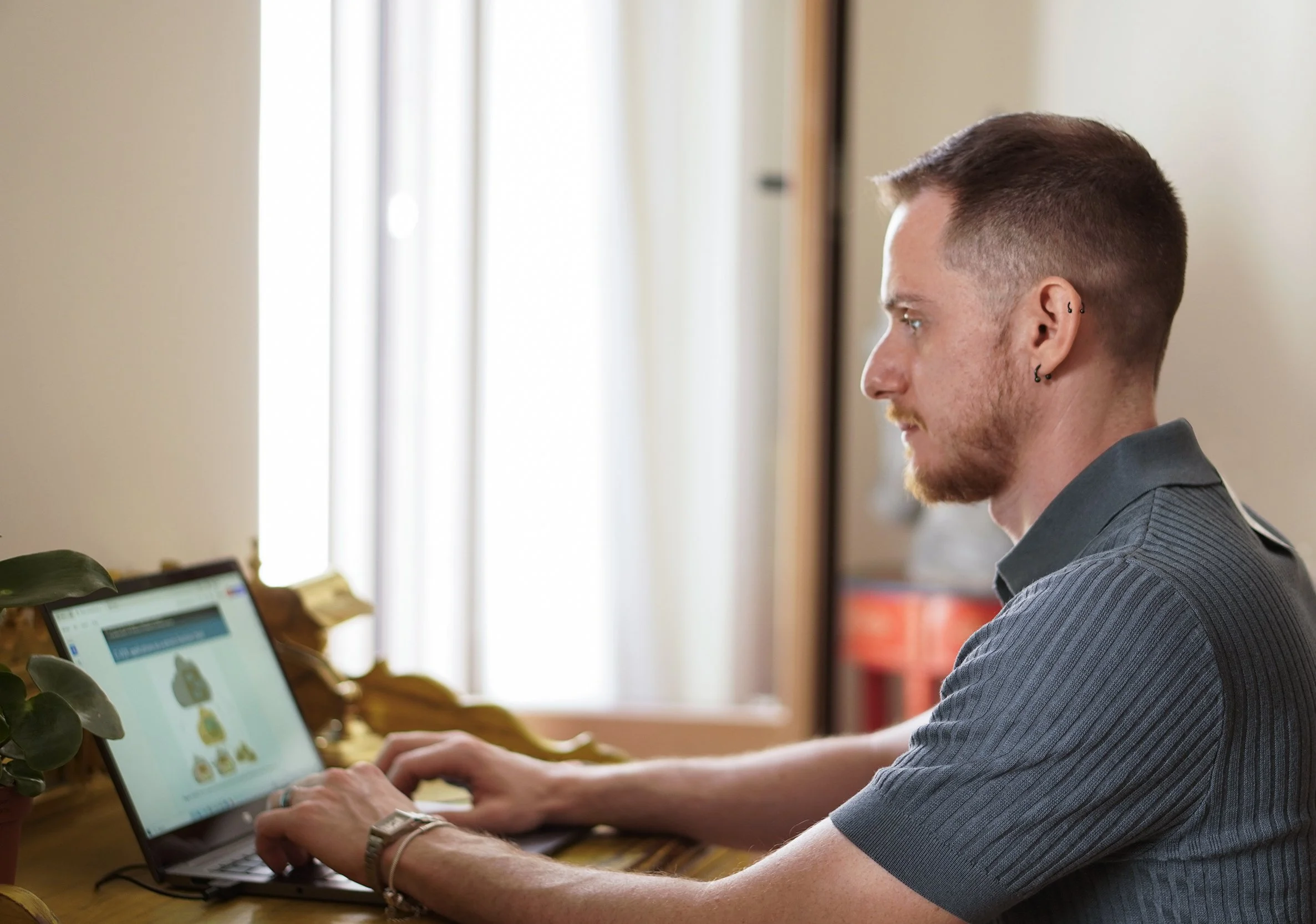 A man with short brown hair and multiple earrings, wearing a gray shirt, is sitting at a wooden desk using a laptop. The laptop screen displays a website or app with images of food. The background features a window with natural light streaming in and beige walls.