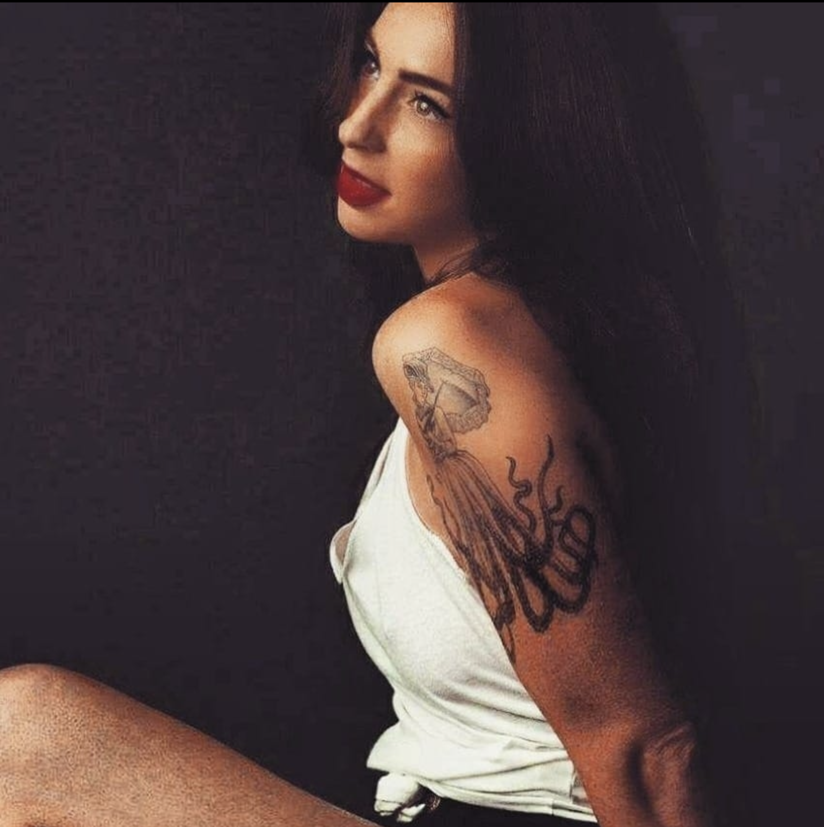 A woman with black hair and fair skin, wearing red lipstick, looking over her shoulder against a black background. She has a tattoo on her right arm.