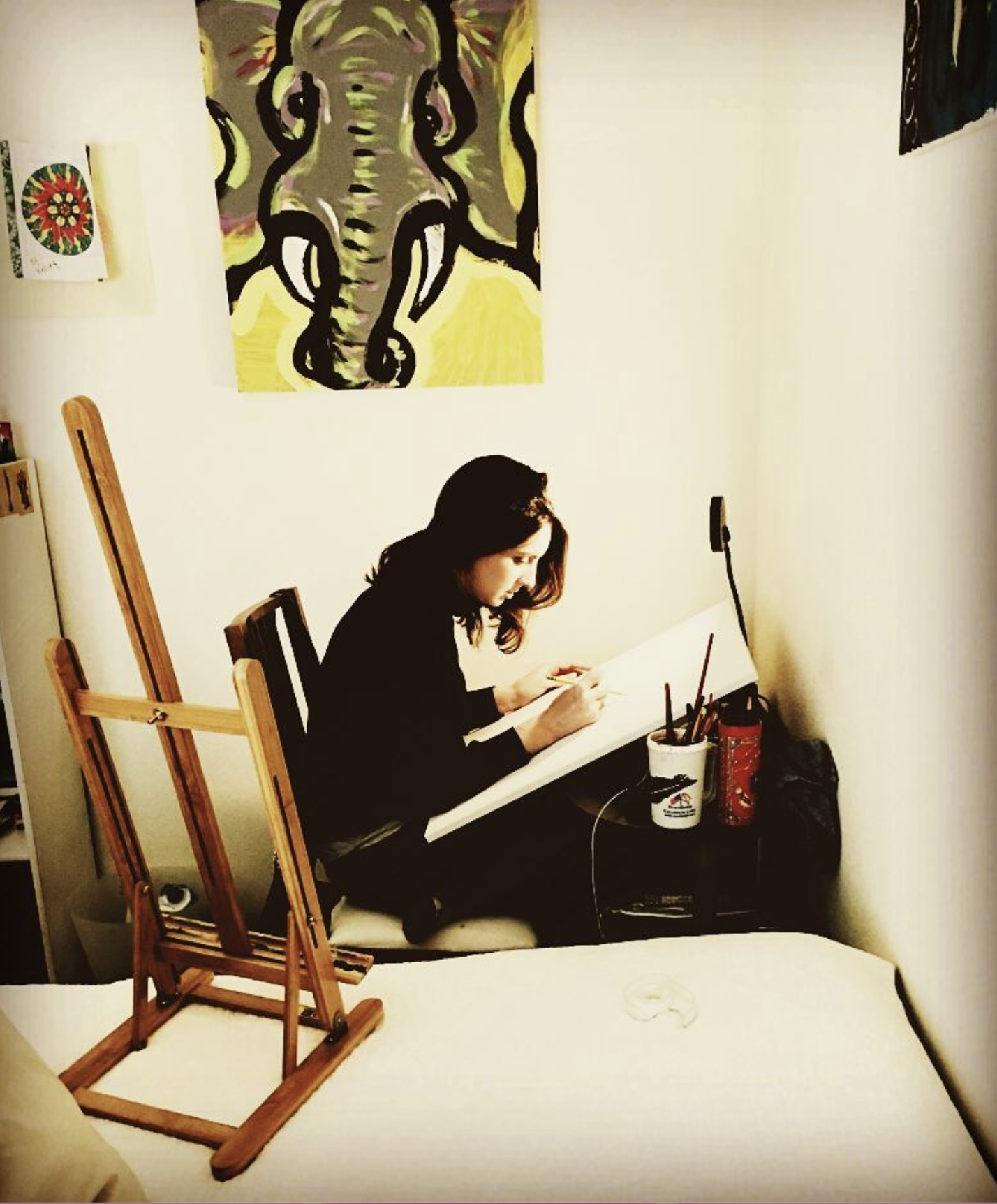 A woman sitting at a table in a room, drawing or painting on a large sheet of paper. On the table, there are containers with brushes and art supplies. An easel is positioned in front of her, and a large abstract painting of an elephant is on the wall behind her.