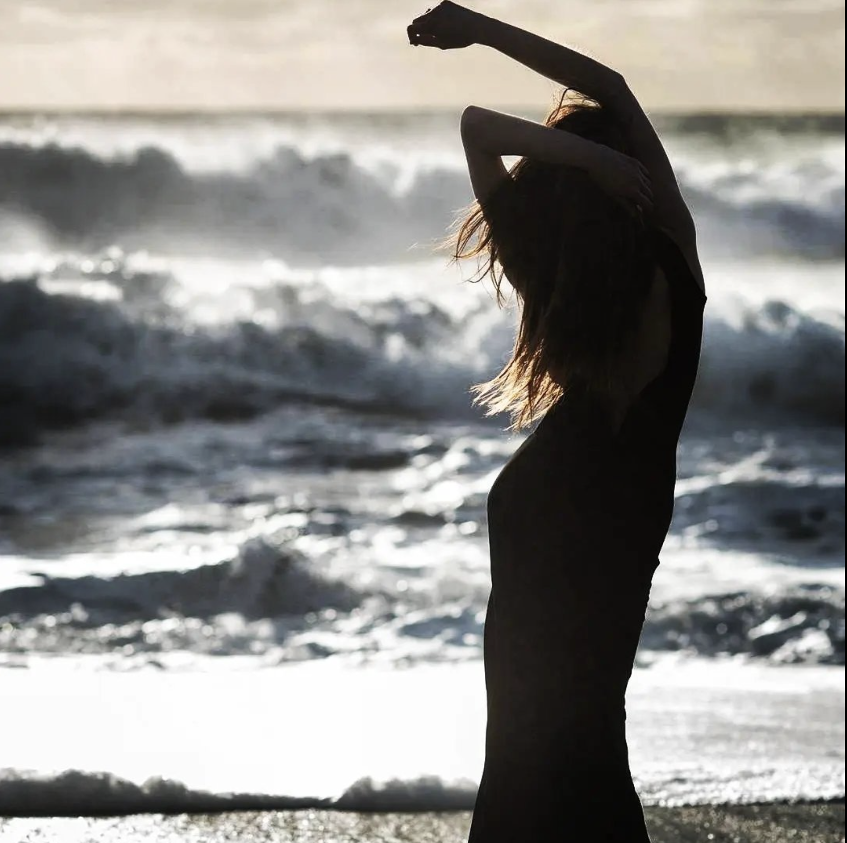 Silhouette of a woman standing on the beach during sunset, with her arms raised and long hair flowing, ocean waves in the background.