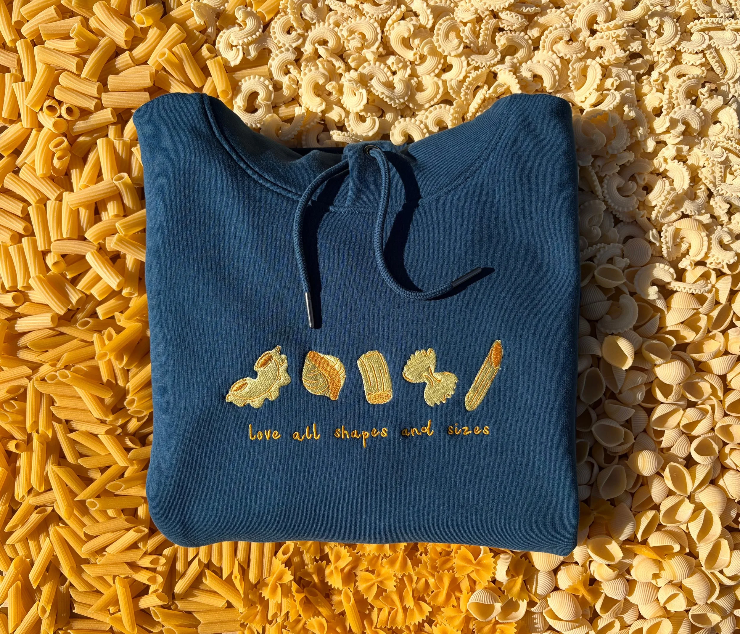 A blue hoodie with embroidered pasta shapes and the phrase 'love all shapes and sizes' on it, placed on a background of various uncooked pasta.