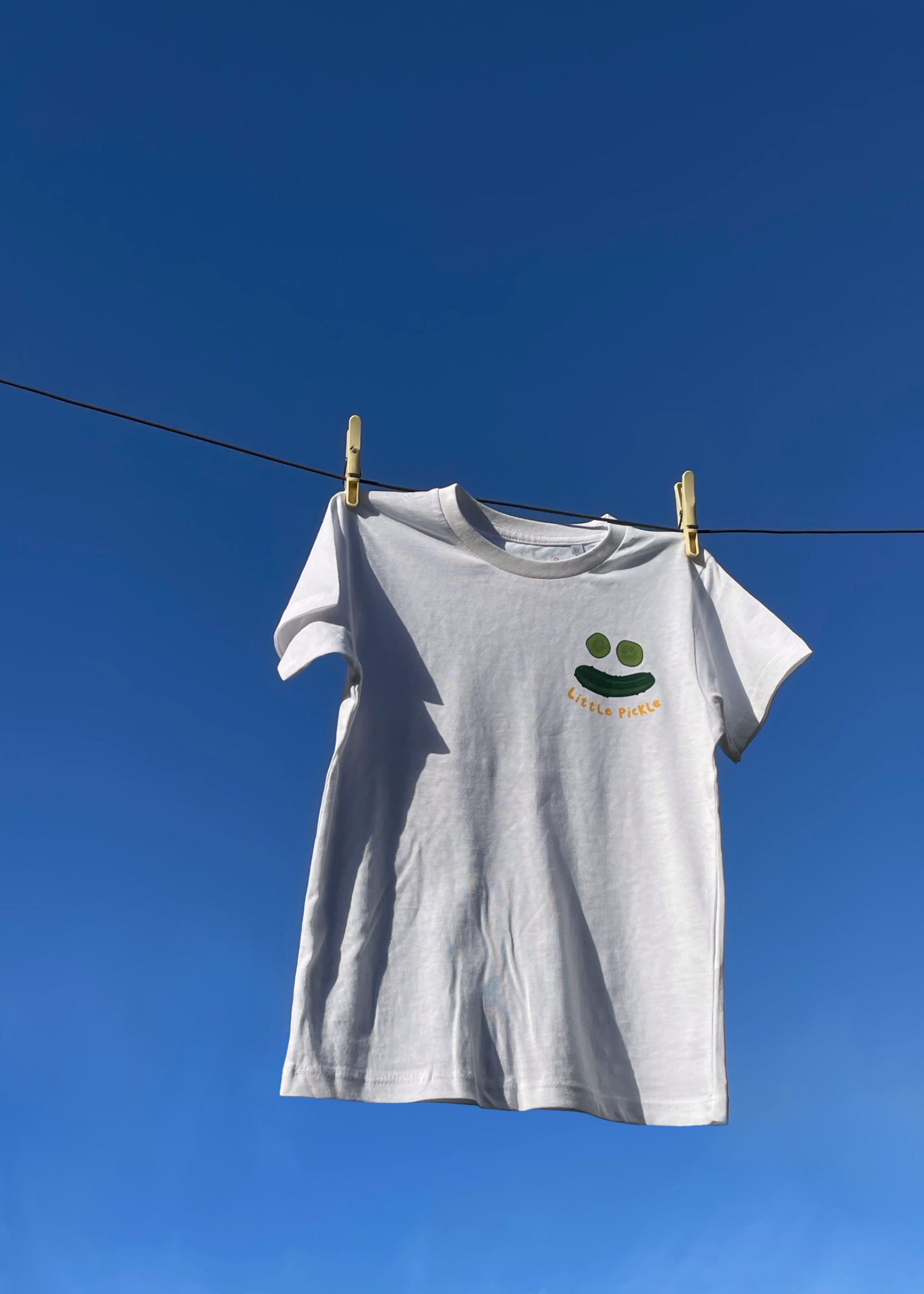 White T-shirt hanging on a clothesline with a drawstring face and the words "Little Pickle" printed on it, against a clear blue sky.
