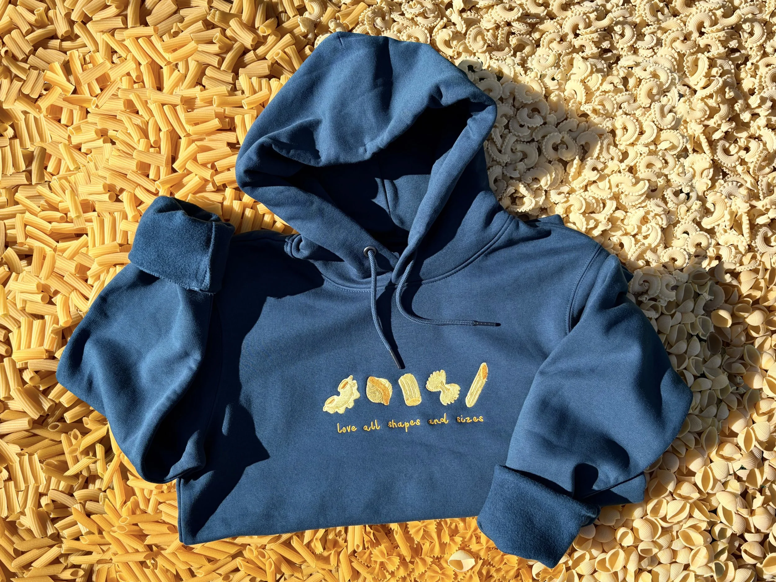 A blue hoodie with yellow embroidered pasta shapes and the text 'love all shapes and sizes' lying on a surface made of yellow and beige pasta.