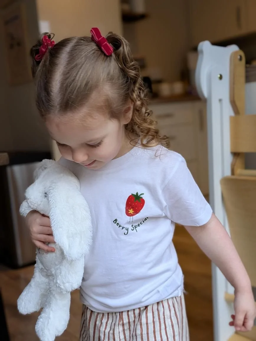 Young girl with curly hair in pigtails held with red hair clips, wearing a white t-shirt with a strawberry graphic and the words 'Berry Special,' holding a white stuffed animal, in a kitchen.