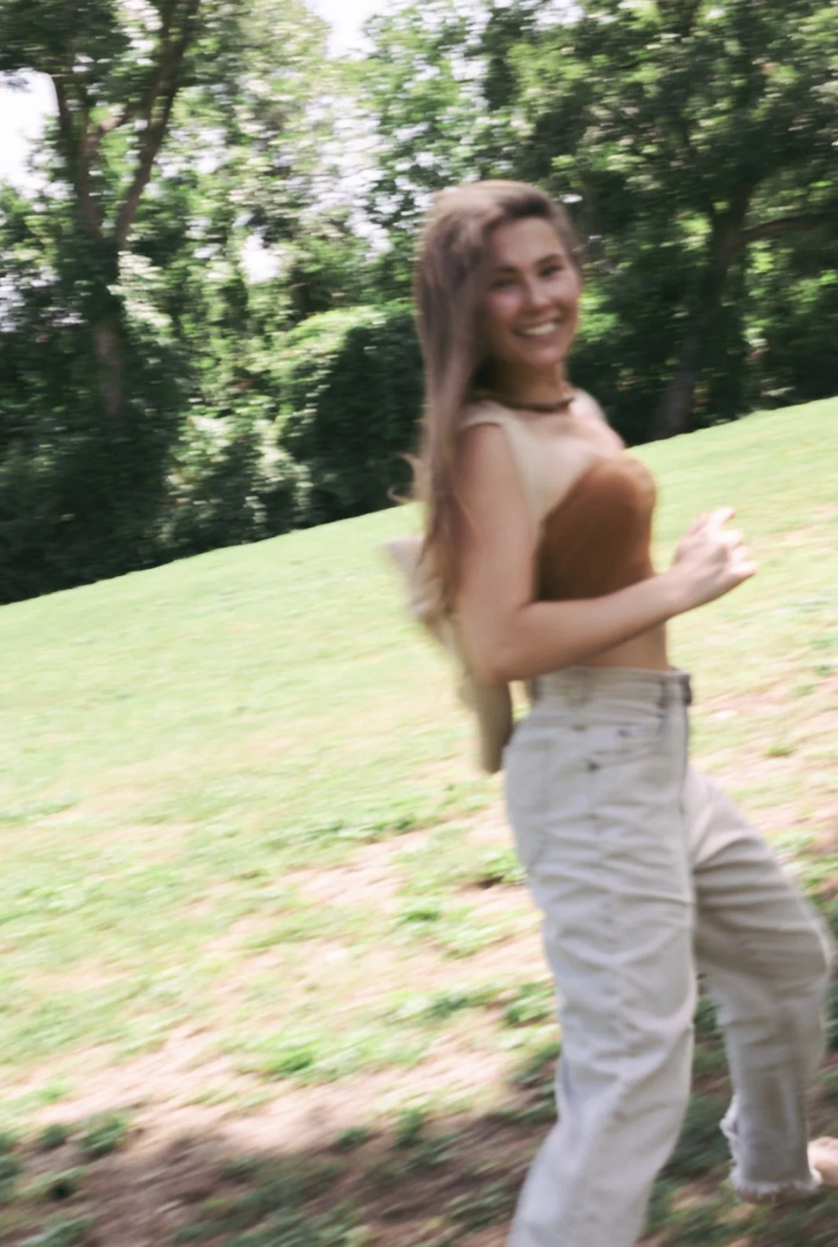 A young woman smiling and running outdoors in a park with green grass and trees in the background.