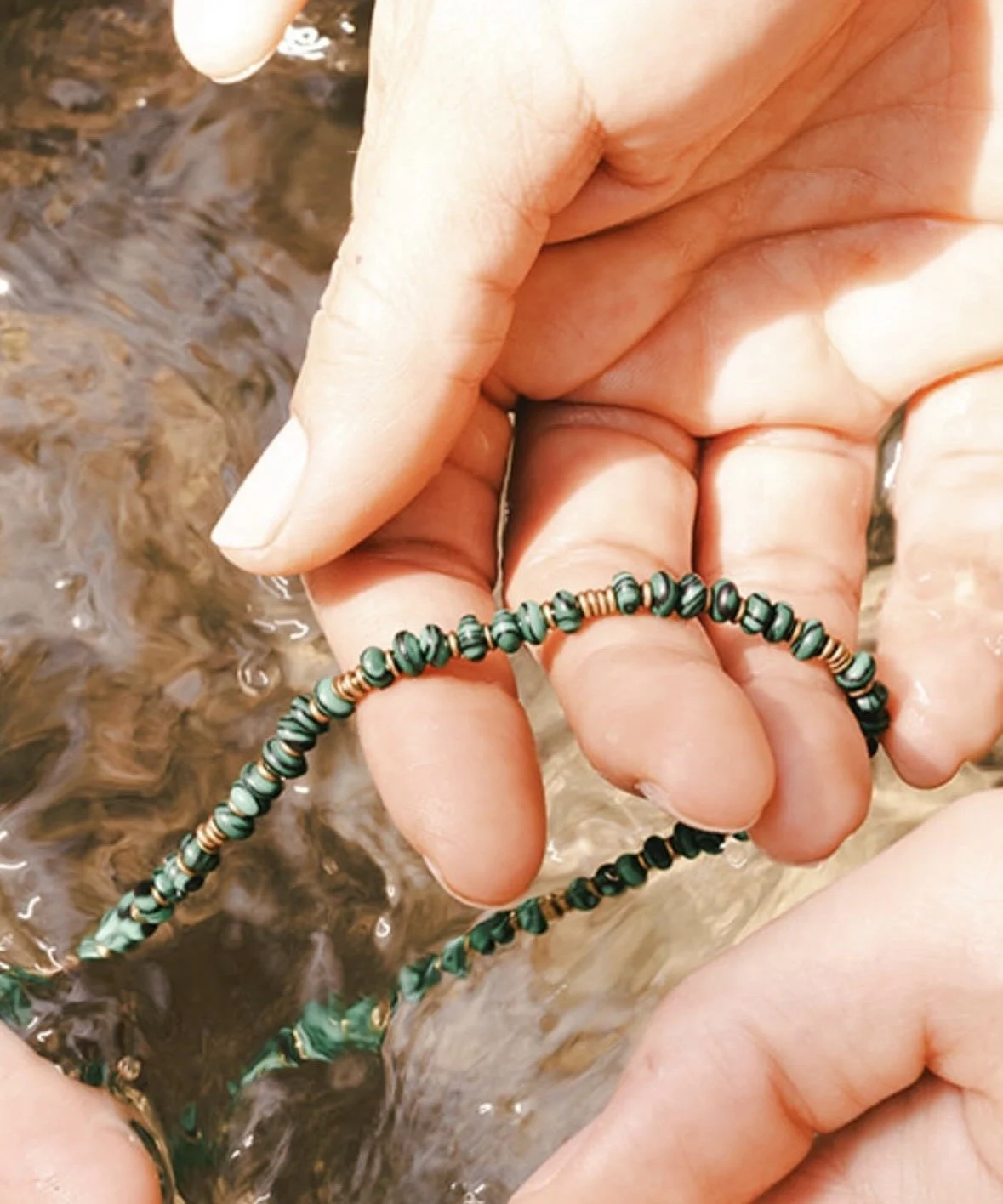 Close-up of hands holding a beaded necklace, with water in the background.