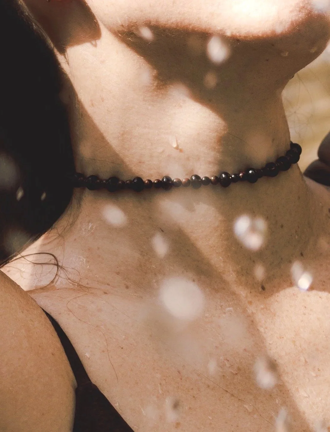 Close-up of a person's collarbone and neck, showing a black beaded necklace, with water droplets on their skin, and soft lighting.