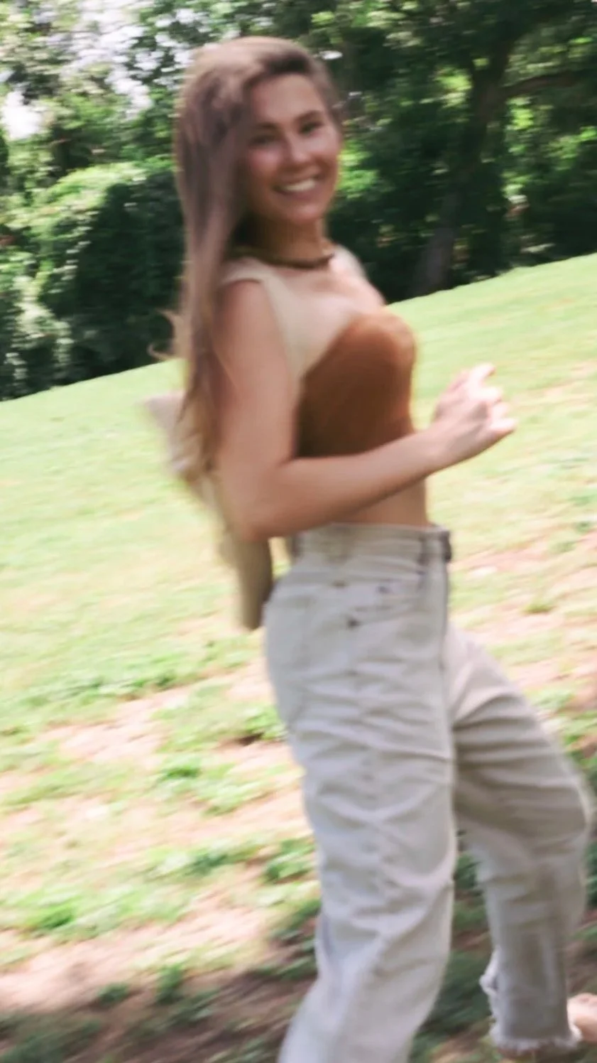 A young woman with long hair smiling and walking outdoors in a park or garden area, wearing a sleeveless top and white pants.