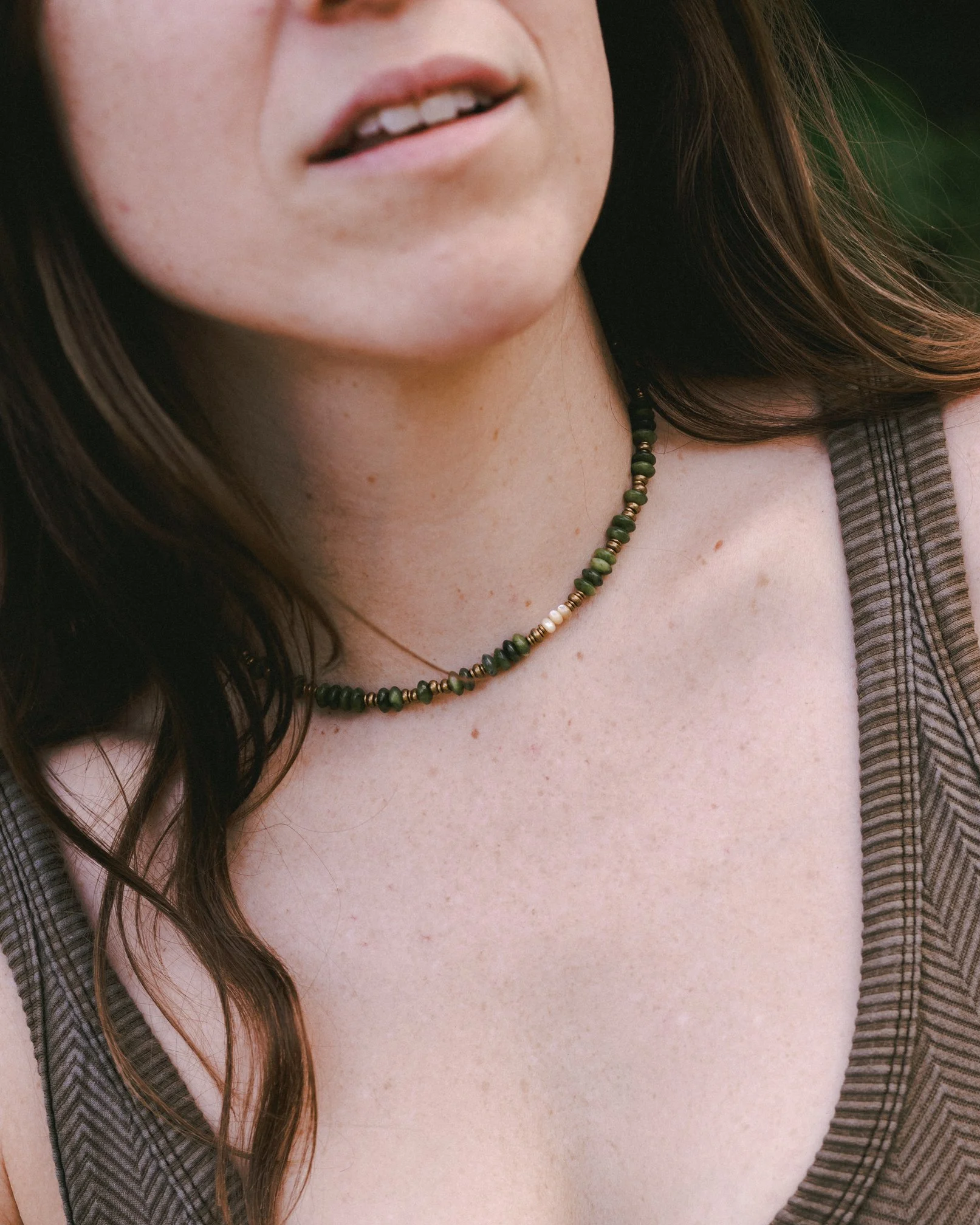 A close-up of a woman's face showing her lips and part of her mouth, with long brown hair and wearing a beaded necklace and a striped shirt.