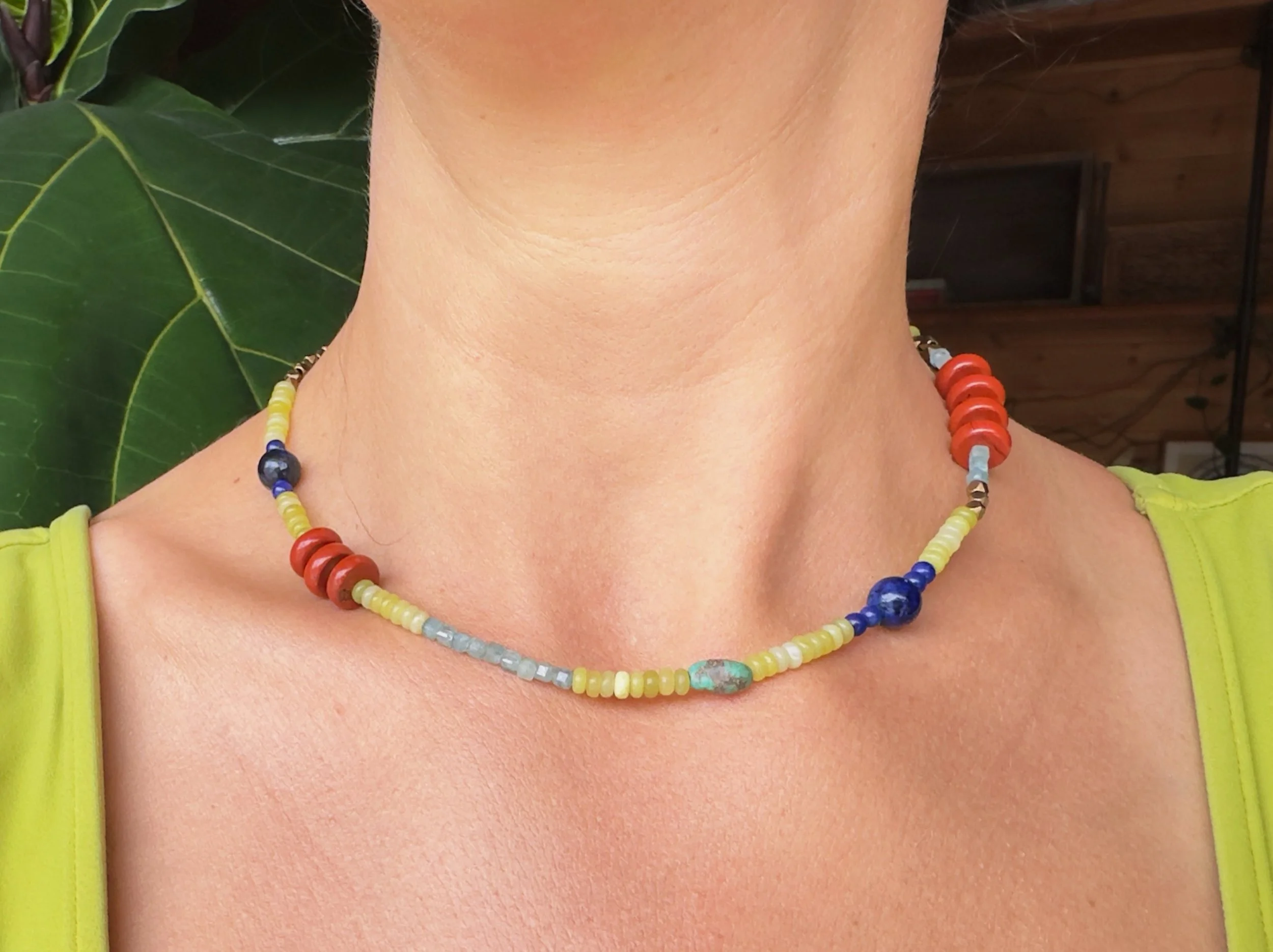 Close-up of a person's neck and collarbone wearing a colorful beaded necklace; to the left, a green leafy plant is visible.