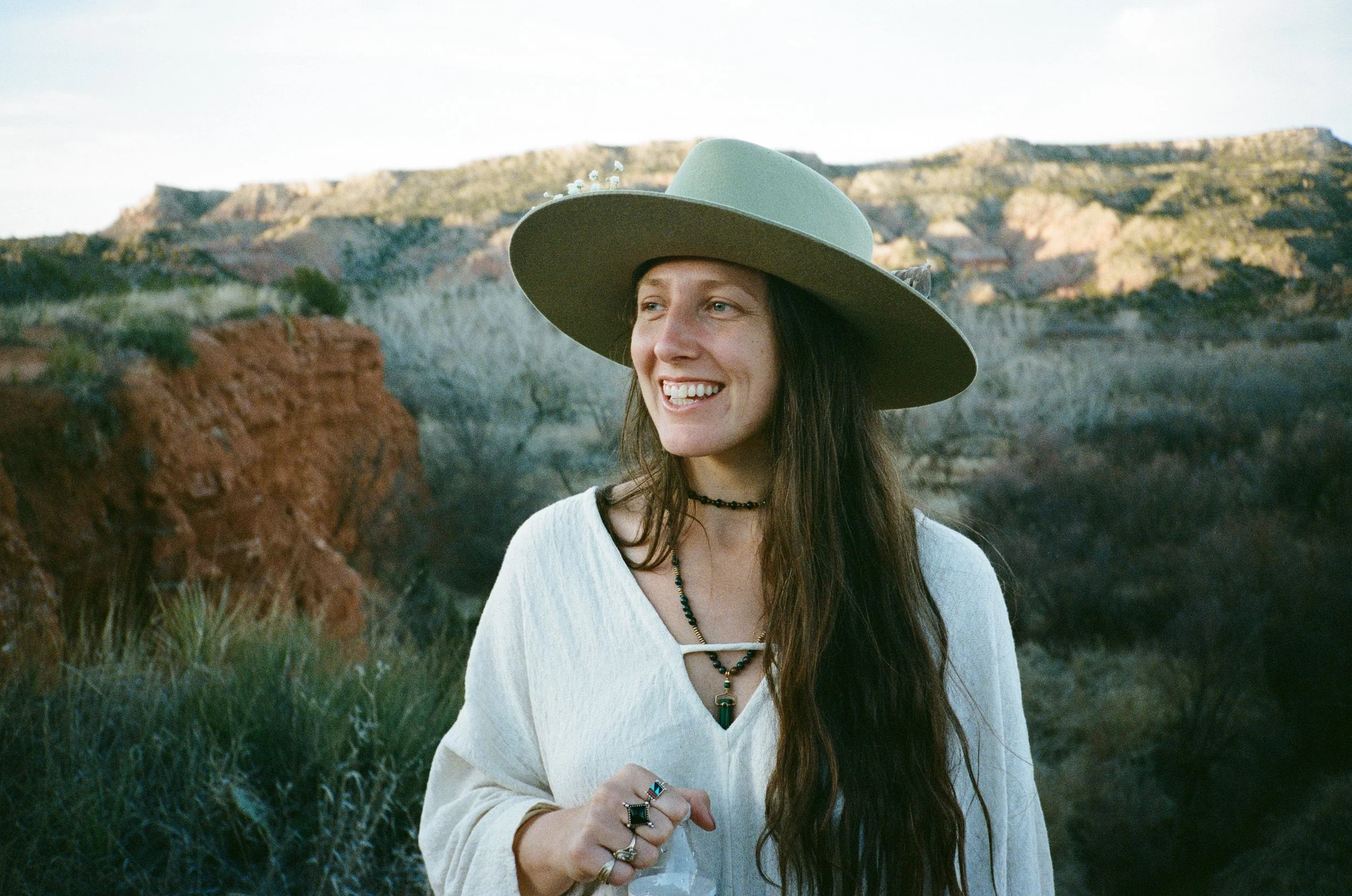 A smiling woman wearing a wide-brimmed hat, a white top, and layered jewelry, standing outdoors with red rocks and cliffs in the background.