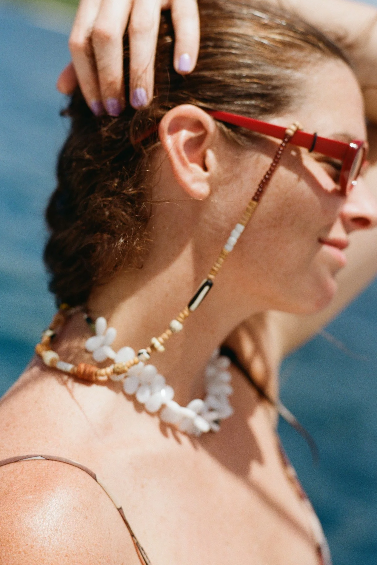 A woman with brown hair, wearing red sunglasses and a black strap on her shoulder, is holding her hair back with her hand. She is wearing a necklace made of white shells and beads and a beaded sunglasses holder.