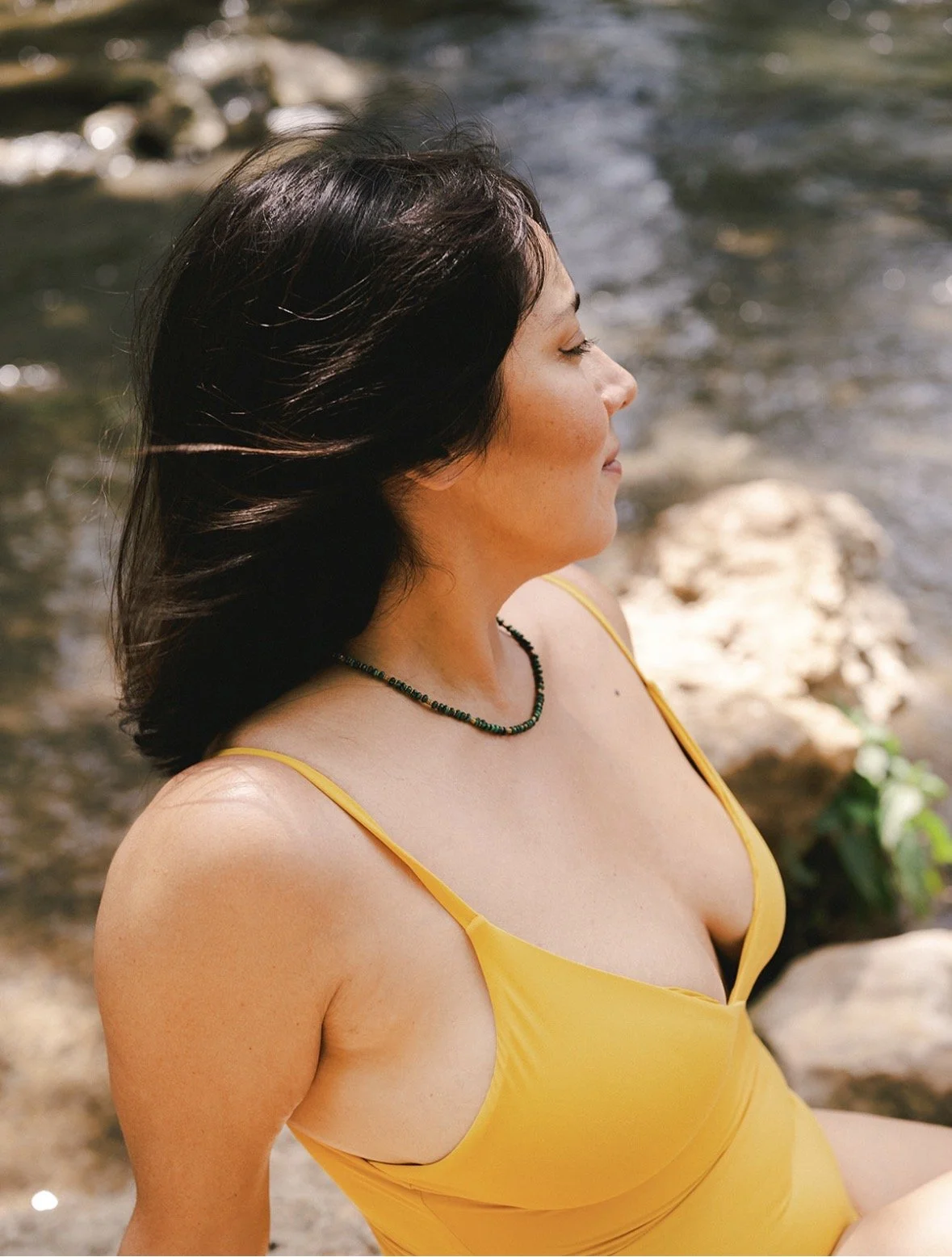 A woman with dark hair, wearing a yellow spaghetti swimsuit and a green beaded necklace, sitting outdoors near a stream with rocks and greenery in the background.
