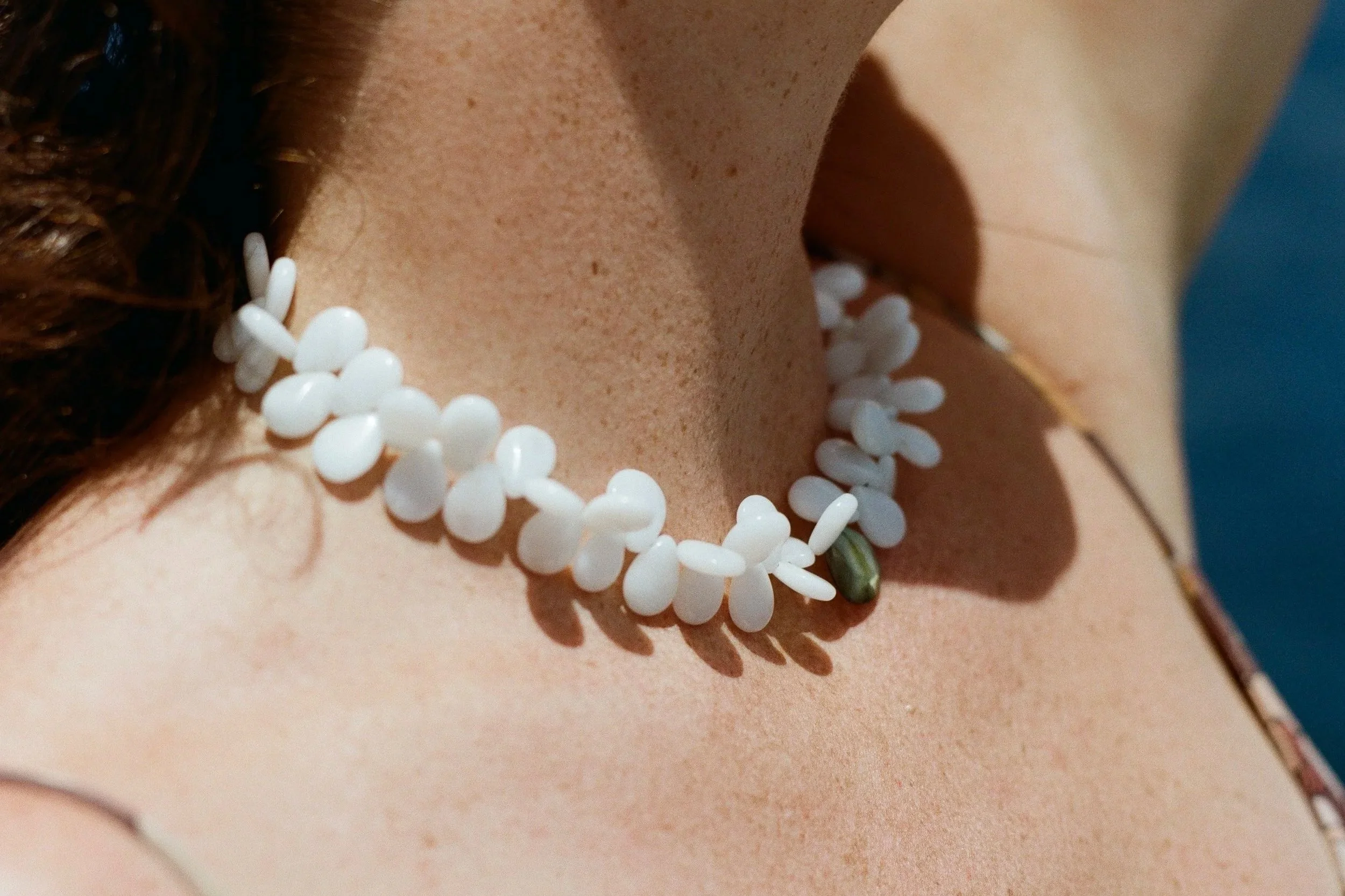 Close-up of a woman's upper chest and neck with white beads necklace, with some brown hair visible on the left side and a blue background on the right.