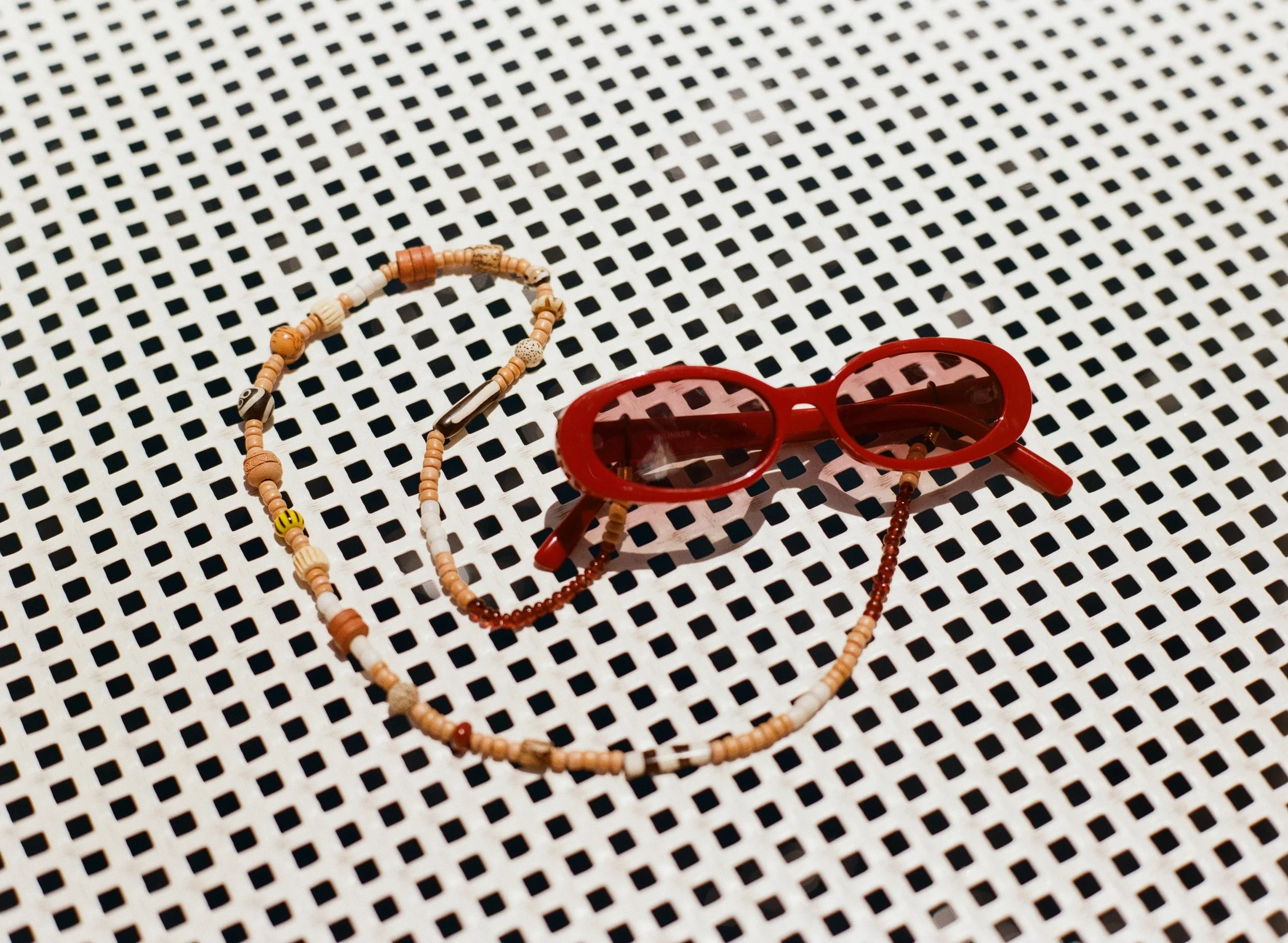 A pair of red sunglasses and a beaded necklace on a black and white checkered surface.