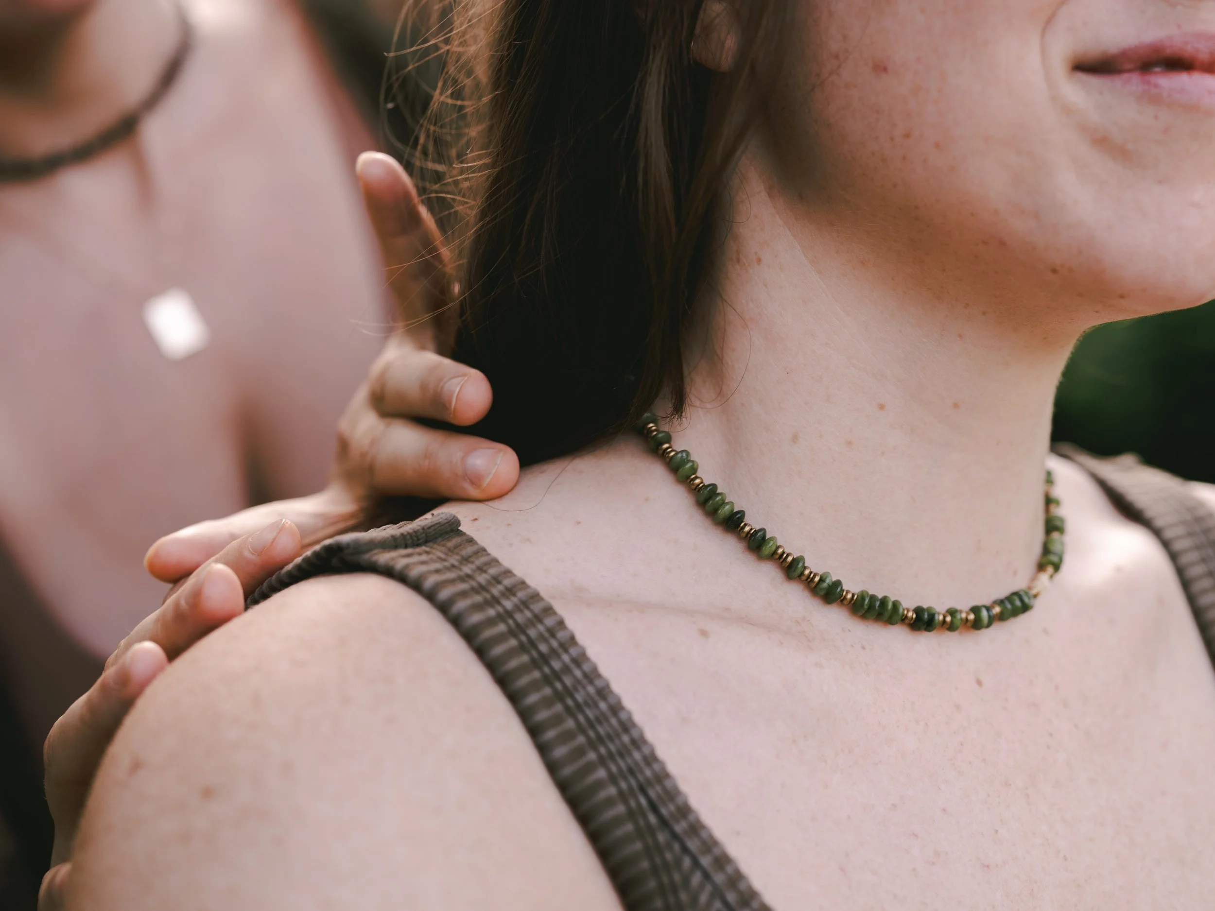 A close-up of a person receiving a shoulder massage from another person outdoors. The individual has freckles and is wearing a beaded necklace. The masseuse's hand is visible on the person's shoulder.