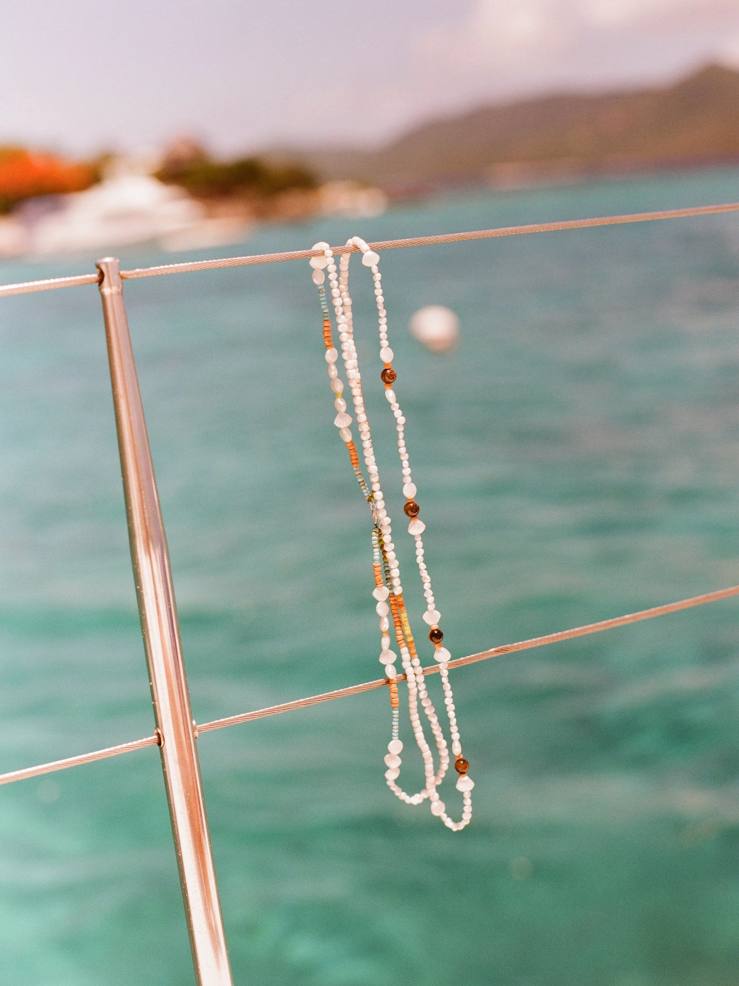 Beaded necklaces hang on a metal railing over turquoise water, with an out-of-focus shoreline and mountains in the background.