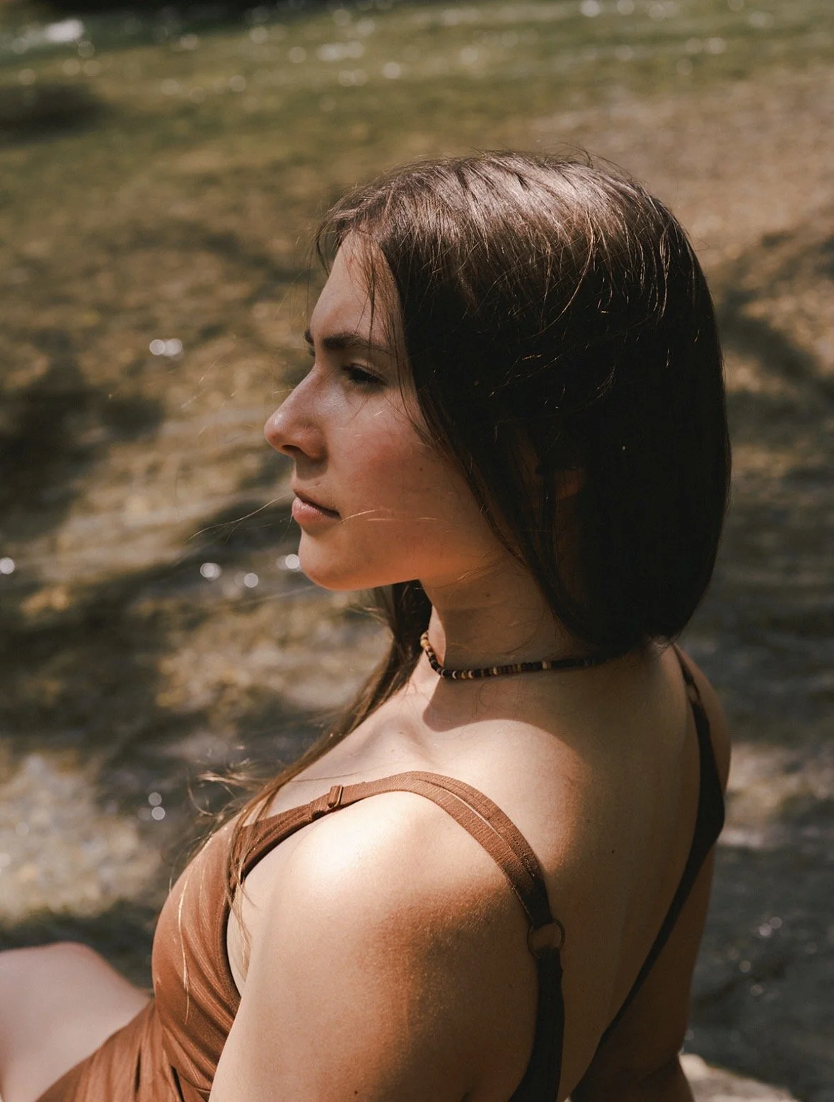 A young woman with brown hair sitting by a body of water, looking to the left, wearing a brown dress and a beaded necklace.