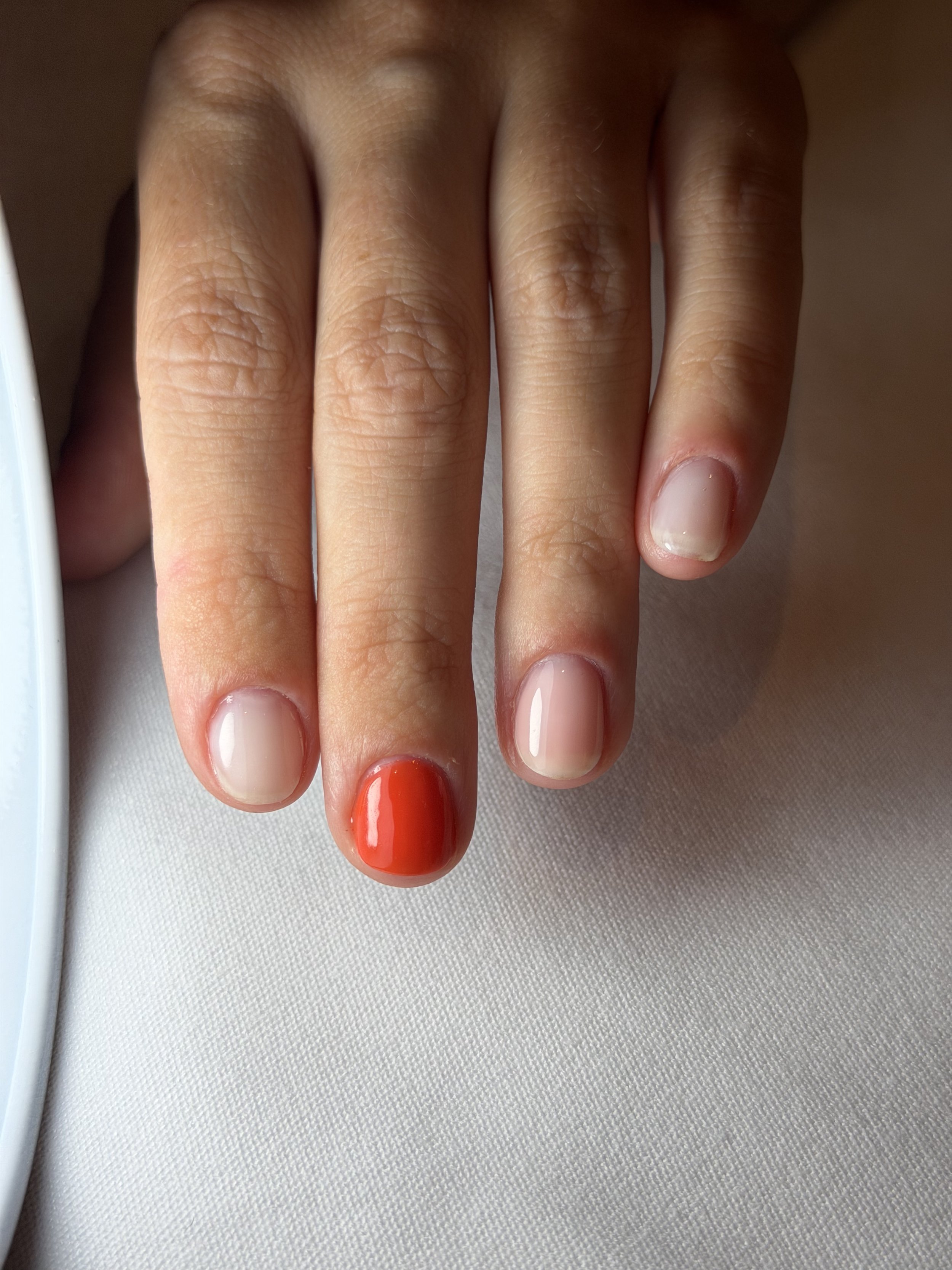 Close-up of a hand with short, natural nails, with the thumb painted in orange nail polish, against a white surface.