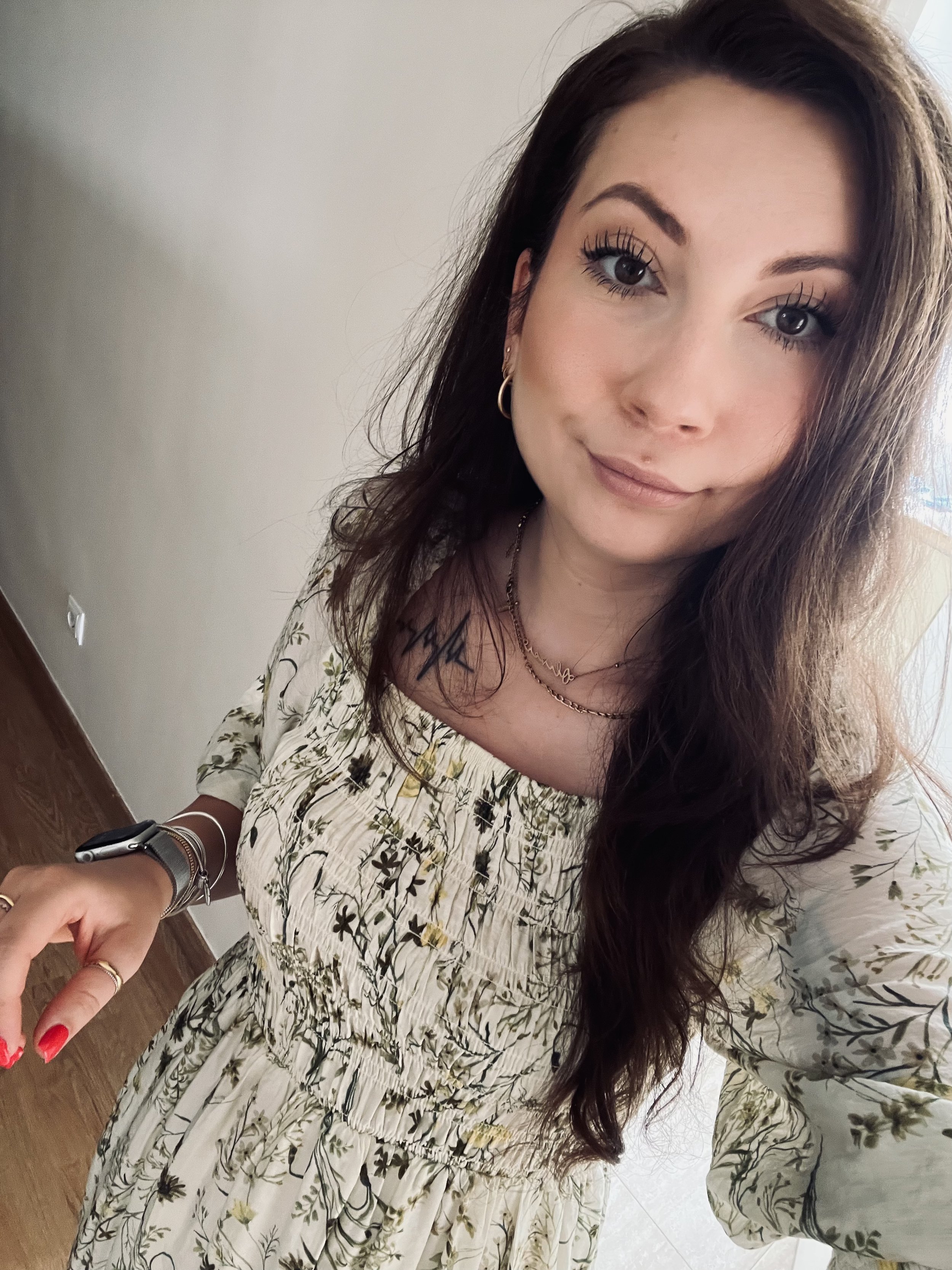 A woman with long wavy brown hair and light skin taking a selfie indoors. She's wearing a floral dress, jewelry, and has makeup on.