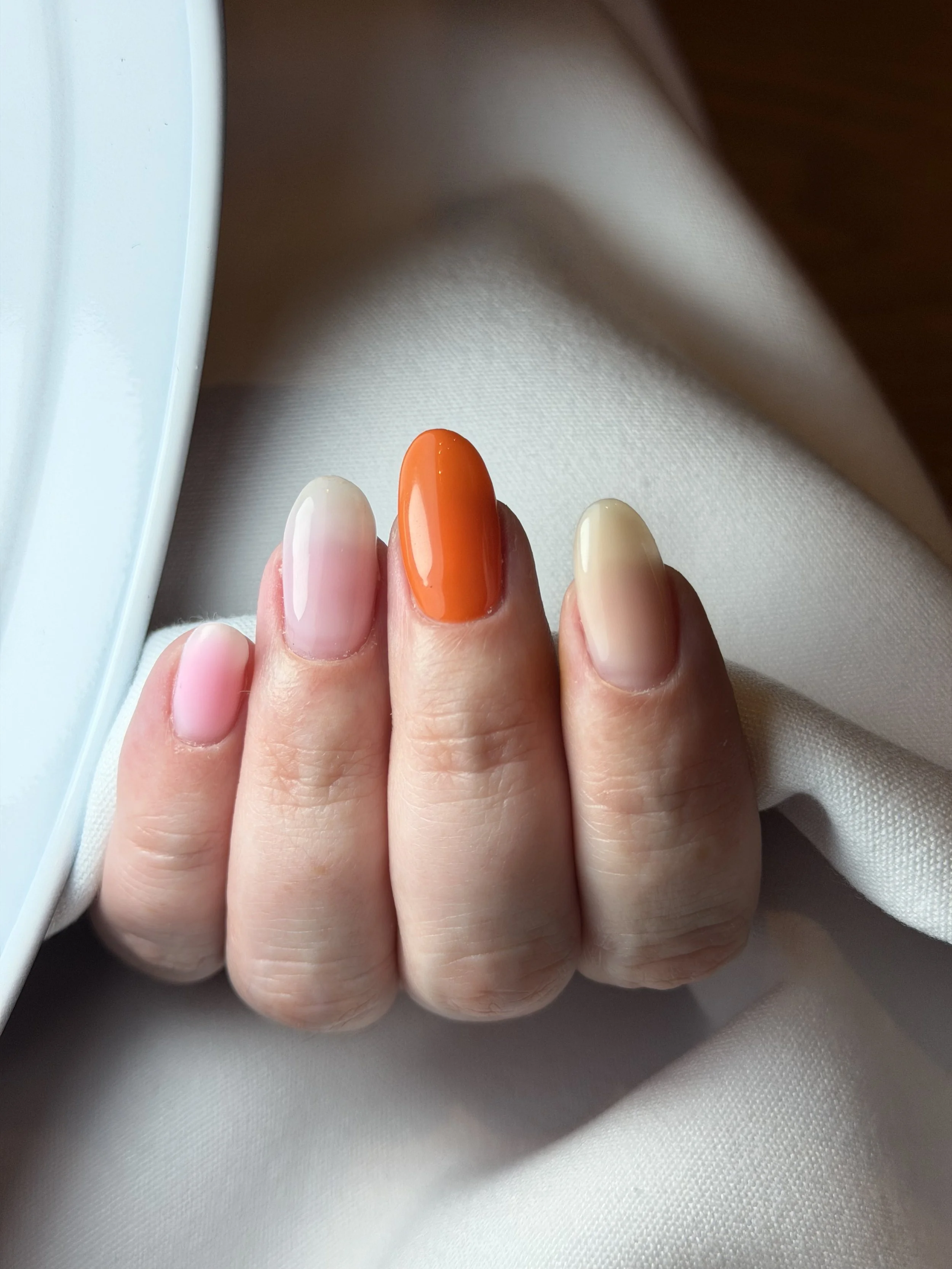 Close-up of a person's hand showing four fingers with different nail colors: two with natural nails, one painted bright orange, and one painted beige, resting on white fabric near a window.