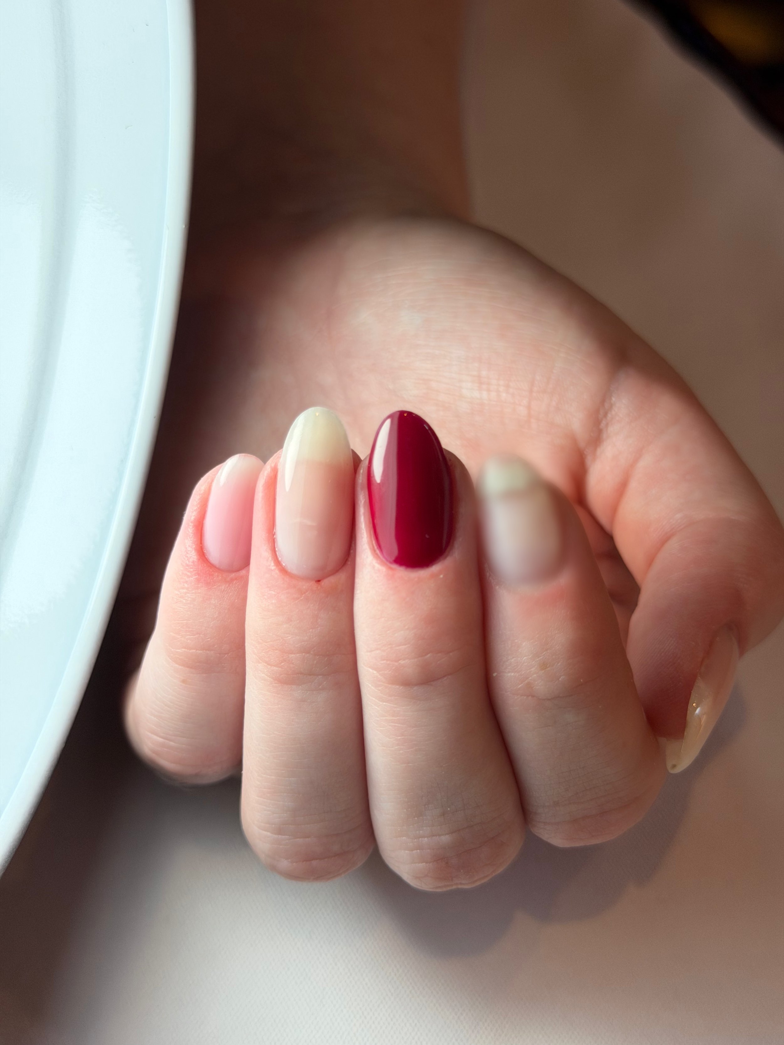Close-up of a hand with manicured fingernails, some painted in a dark red color and others in a light nude color, near a window with natural light.