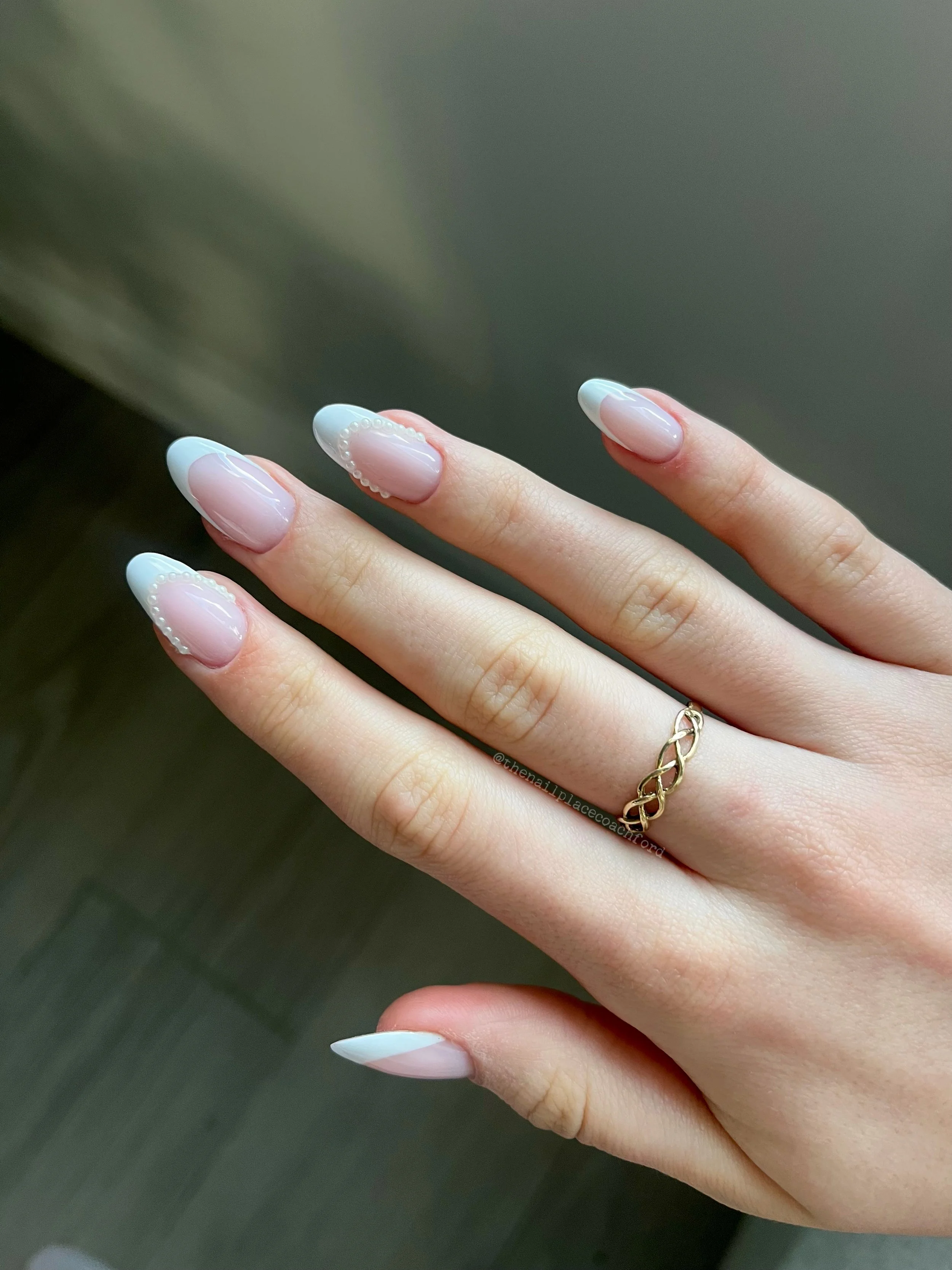 Hand with manicured nails featuring a French tip design and a gold chain ring, against a neutral background.