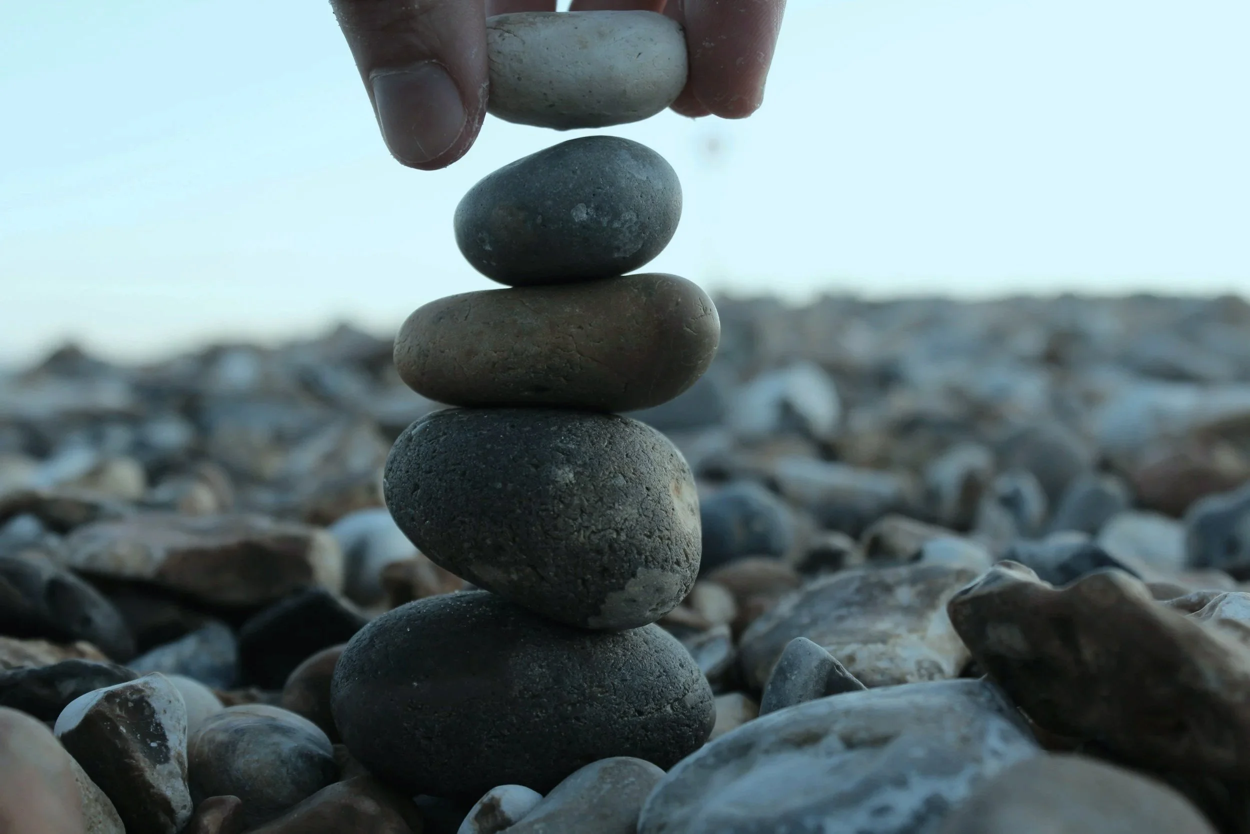 A close-up of a human hand stacking five smooth, rounded stones on top of each other on a rocky beach with a blurred horizon in the background.