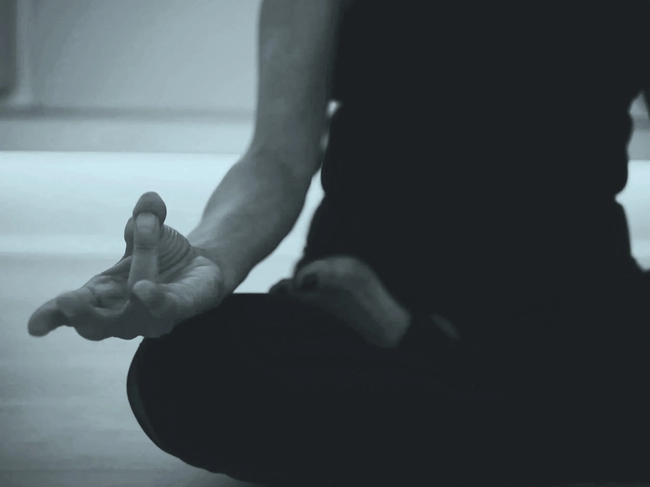 Person sitting cross-legged with their arm extended forward, hand open in a gesture, in a dimly lit room.