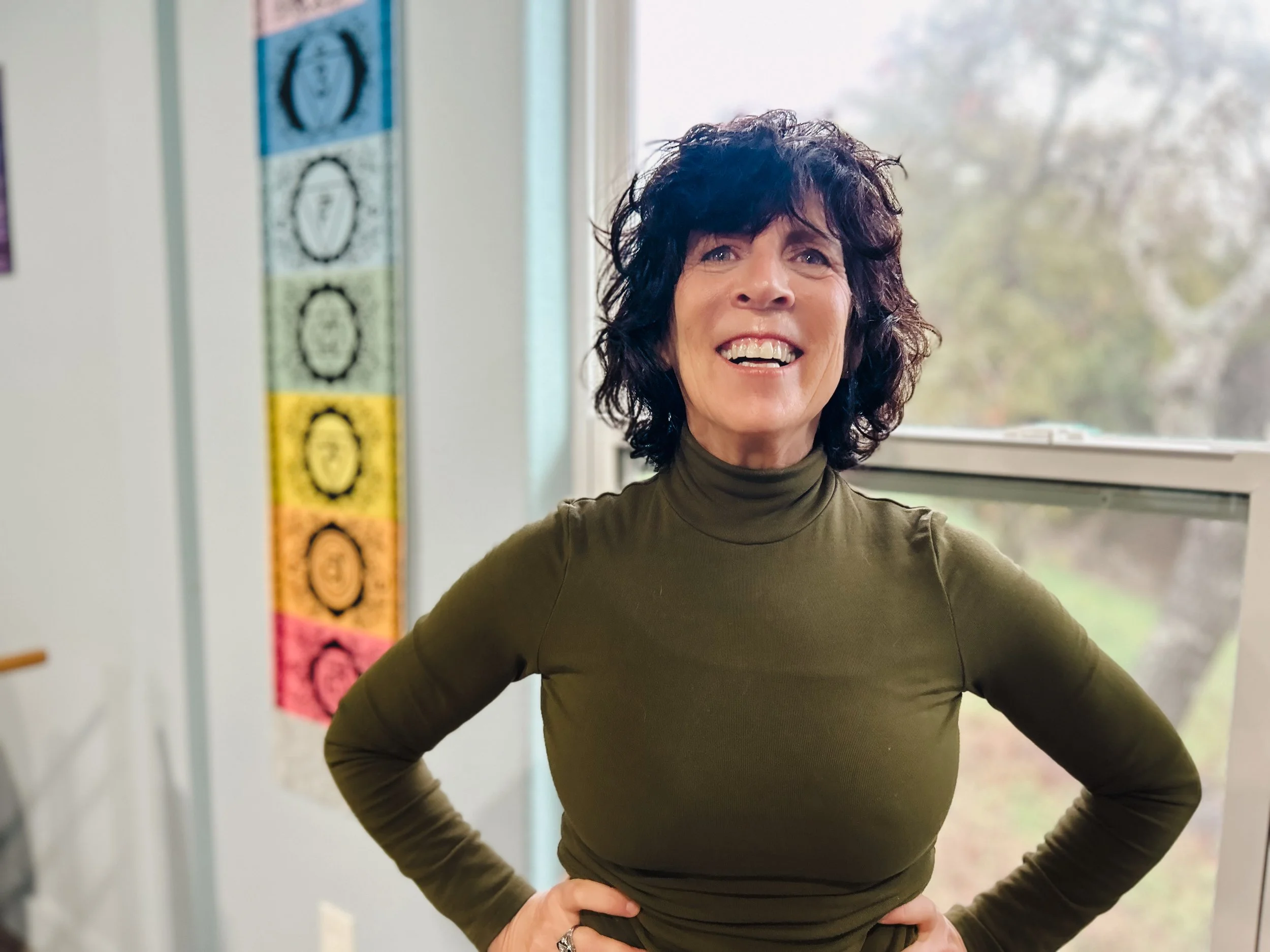 A woman with curly dark hair wearing a green turtleneck shirt, smiling and standing with her hands on her hips indoors near a window.