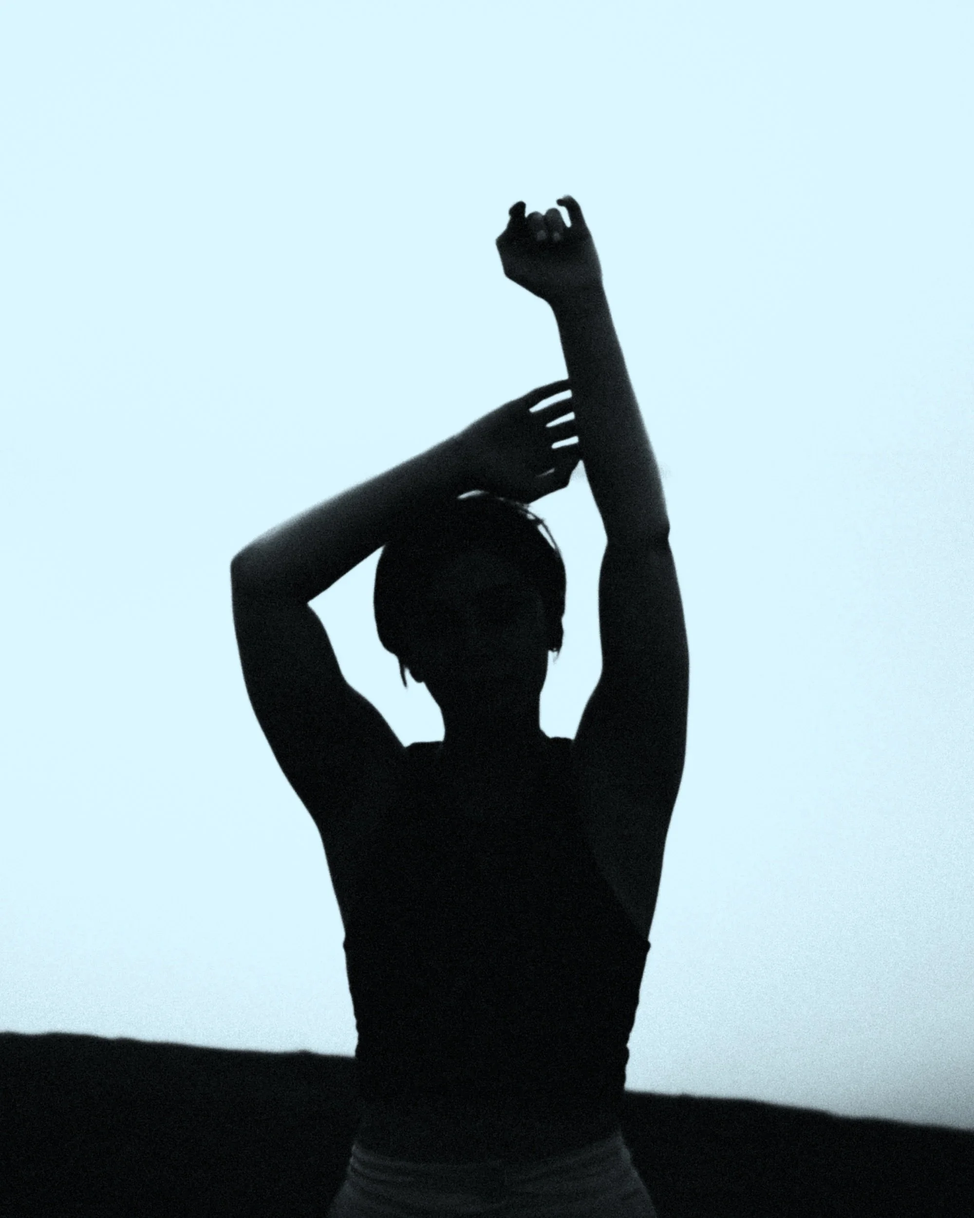 Silhouette of a person with dreadlocks standing outdoors against a light sky, with arms raised, one bent at the elbow and the other extended upward.