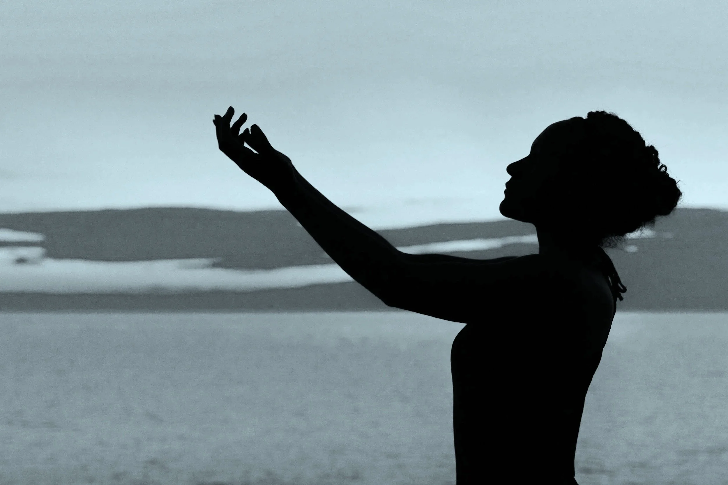 Silhouette of a woman with curly hair reaching out toward the sky at the beach, with waves and horizon in the background.