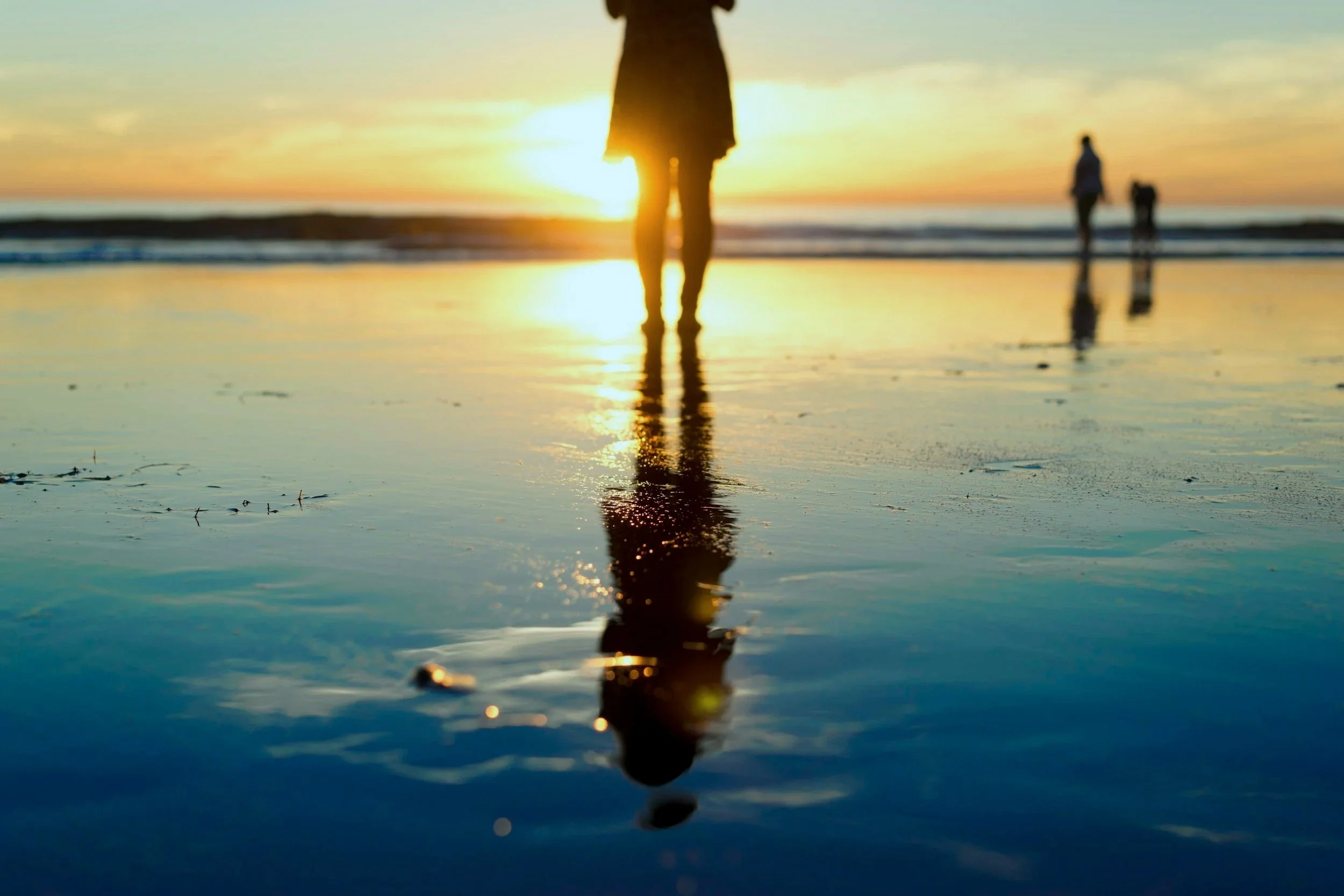 People walking on a beach during sunset, with their reflections visible on the wet sand.