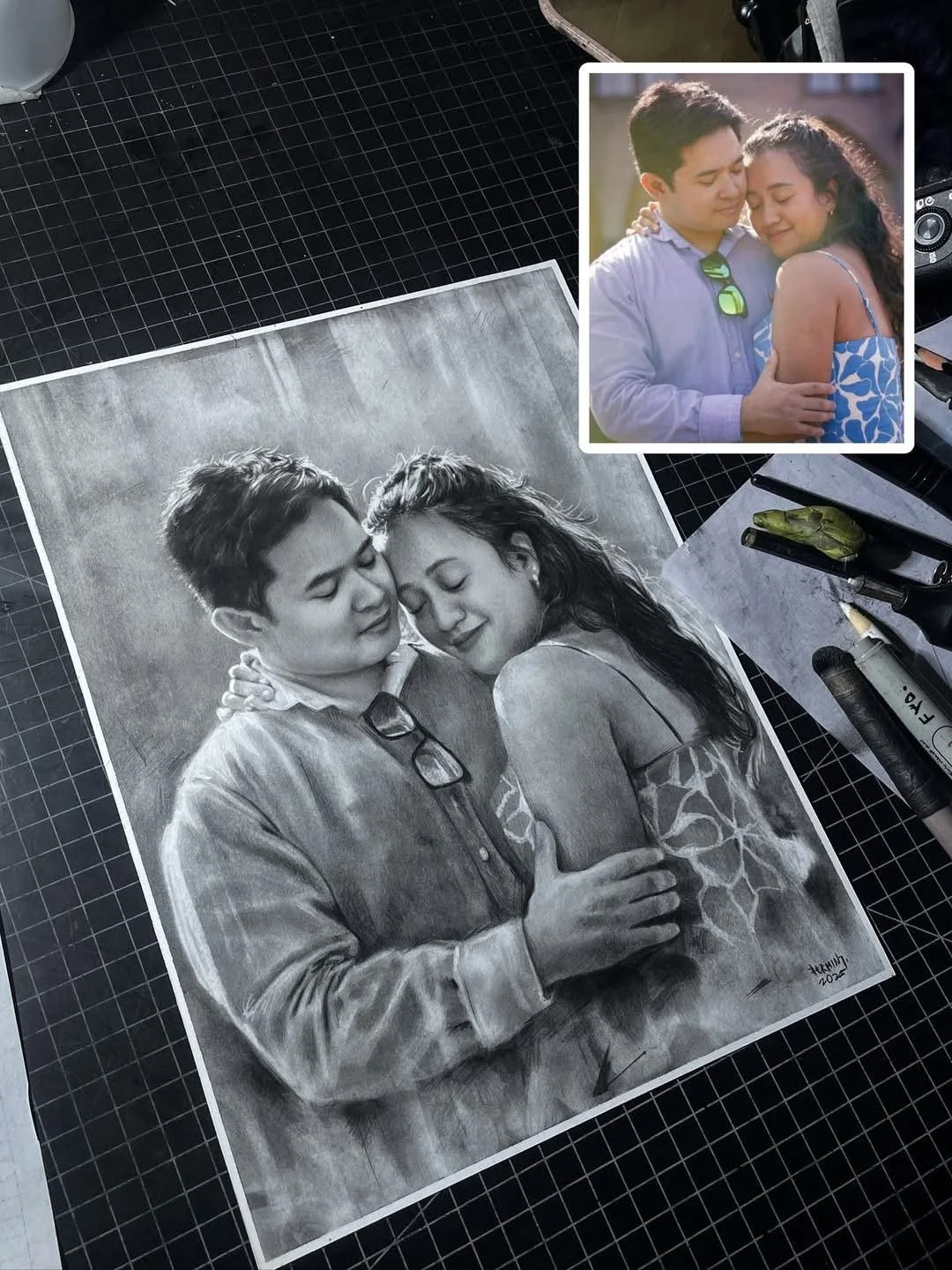 A black-and-white portrait drawing and a colored photograph of a couple embracing, with the drawing showing the couple with their eyes closed, smiling softly, and the woman resting her head on the man's shoulder. The drawing is on a table with drawin