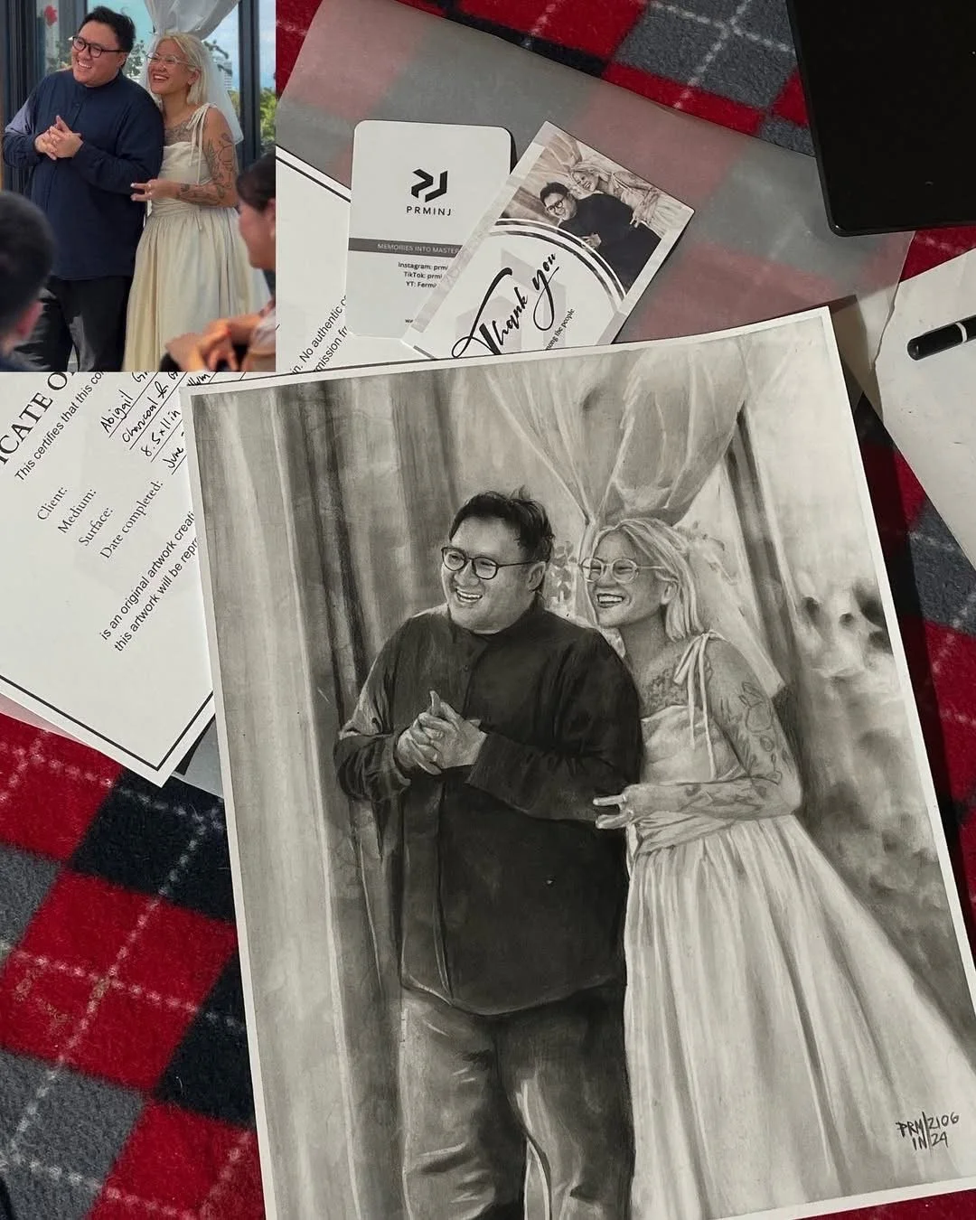 A framed black and white portrait drawing of a smiling man and woman, standing close together. The man is wearing glasses and a dark shirt, while the woman has tattoos on her arms and is wearing a light dress. The background features curtains or drap