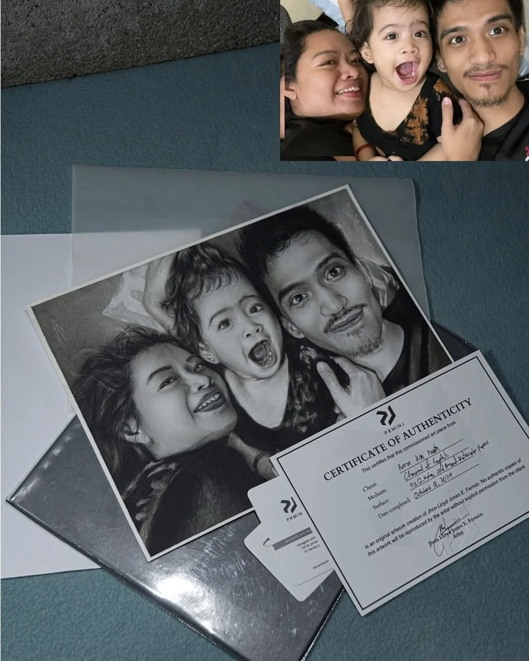 A printed black-and-white photograph showing a woman, a young girl, and a man making playful faces, lying down on a surface. The photo is accompanied by a certificate of authenticity and an ID card, indicating it is an original artwork by artist John