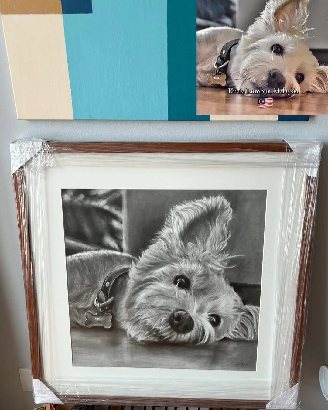 A framed grayscale realistic pencil drawing of a dog lying on the floor, with a close-up of its face and one ear raised. The dog is wearing a collar with a bone-shaped tag, and the scene appears indoors.